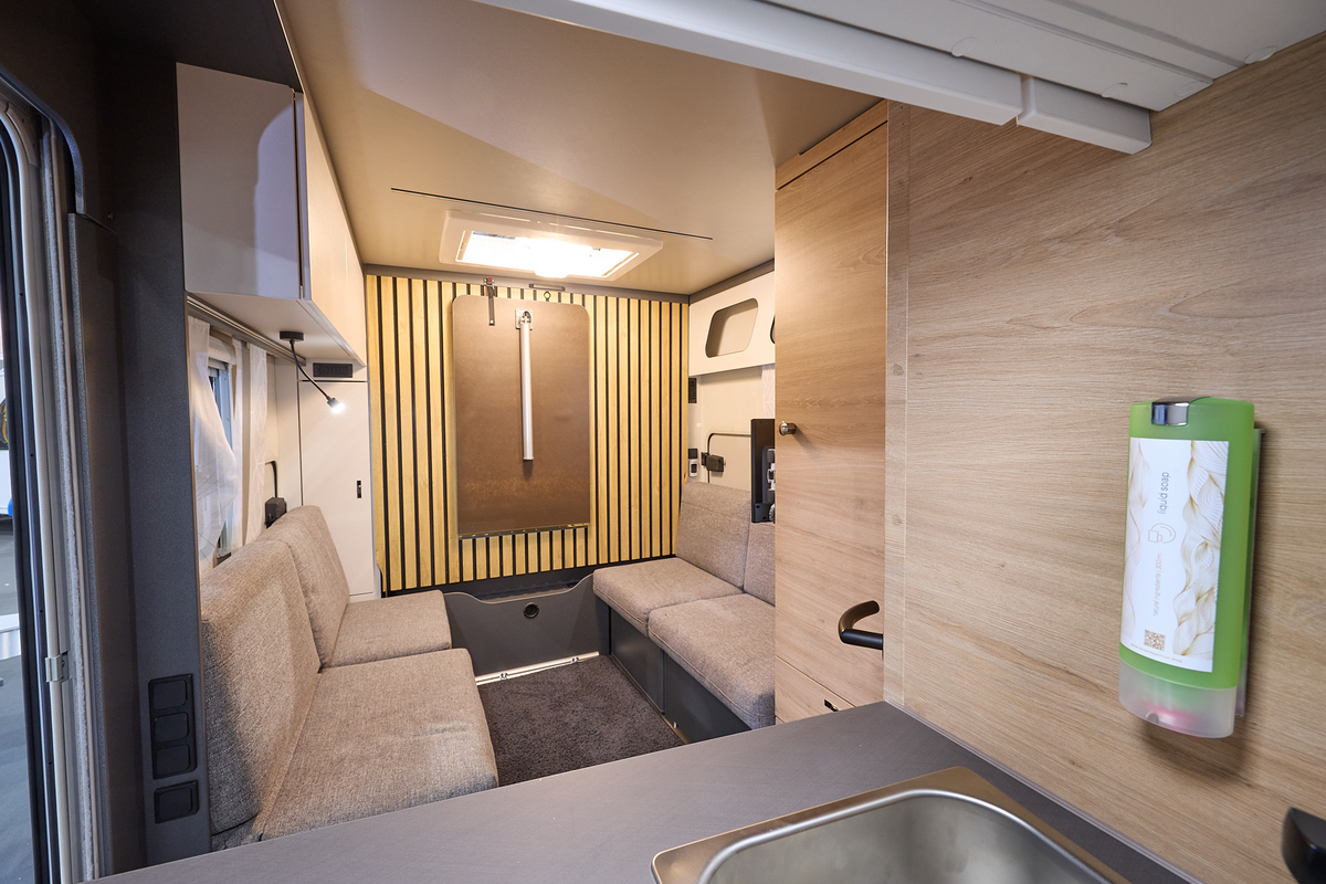 Knaus Yaseo 340 PX's interior is pale wood with beige upholstery.  There is a dispenser with green soap at the front.  To the rear there is a panel of strips of wood which includes a holder for the table which held in this position.