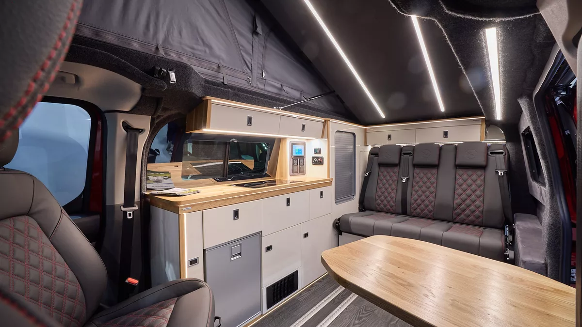 The Landseer Artisan Red Ford Custom 2.0 has a large wooden table and its upholstery is black with red stitching.  The cabinets are cream and pale wood.  The rising roof is fully open.