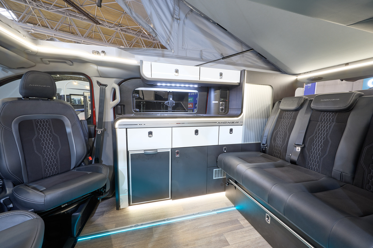 The Swift Monza has wooden flooring with black upholstery.  The cabinets are white and dark blue.  The pop top roof, along with its canvas, is open.