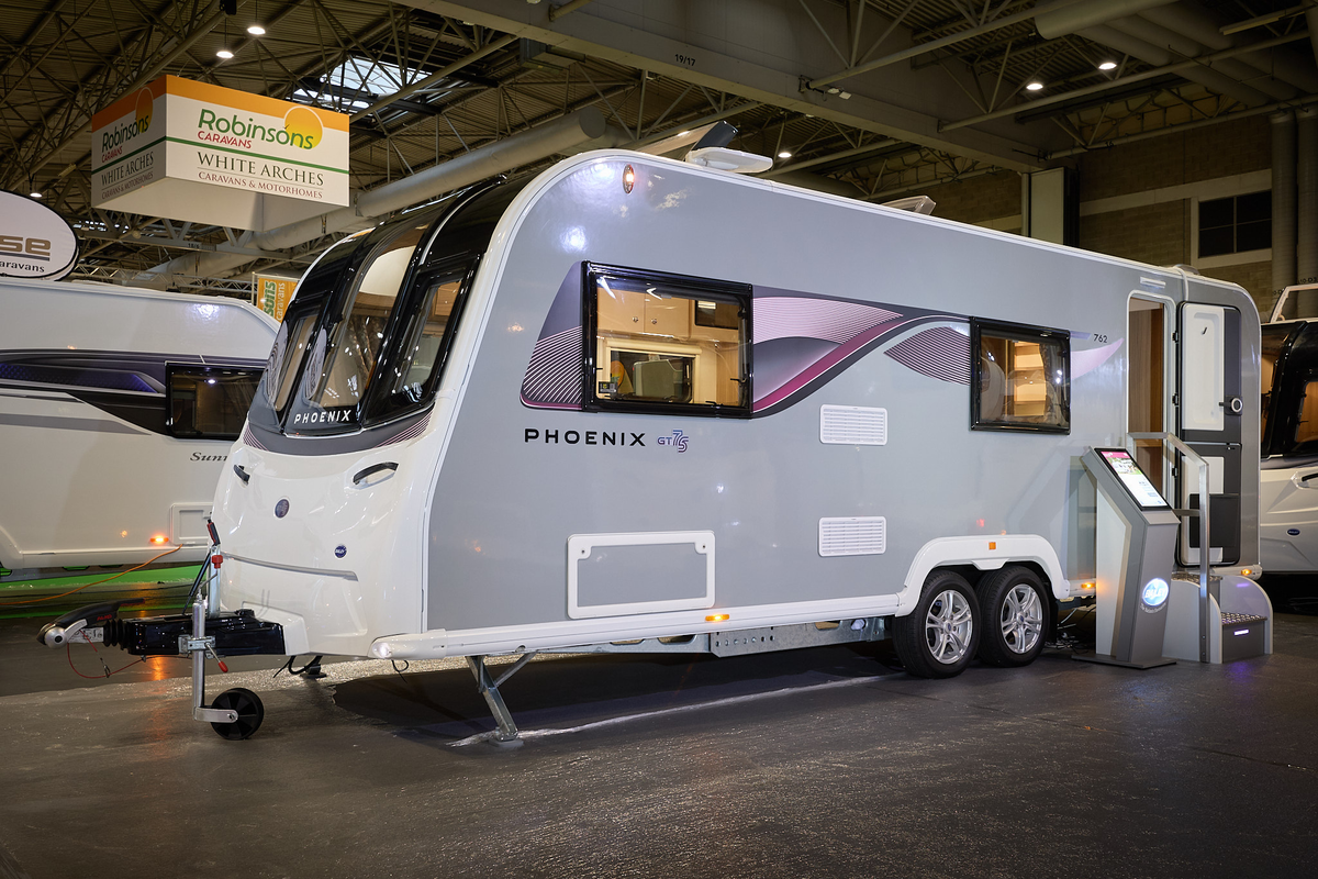 Bailey Phoenix 762 exterior’s grey and white with a large single front window.  The entrance door is open and there are fixed metal steps into its interior.  It is a twin axle. There is an interactive stand next to the steps.