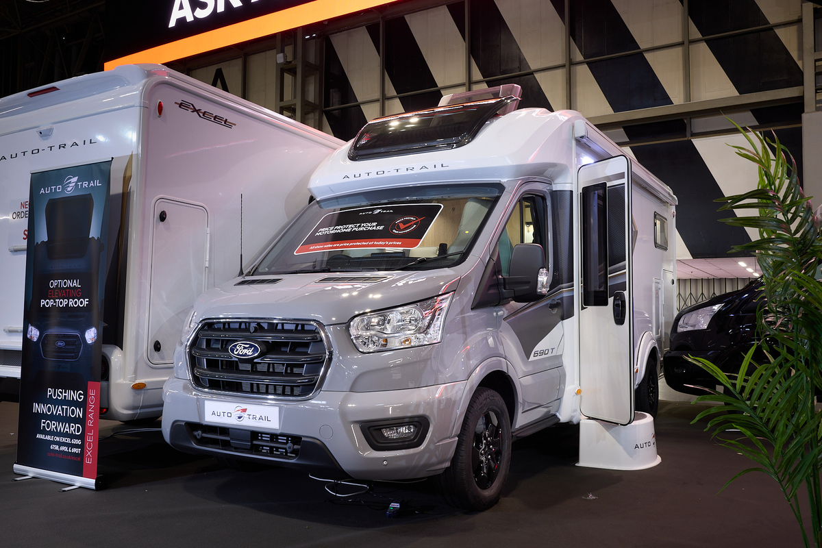 The Auto-Trail Excel 690T has a silver cab and white body with grey decals.  There is a show offer on its windscreen in red.  There is a white step by the opened habitation door.  It has a large sunroof and its roof light is open.