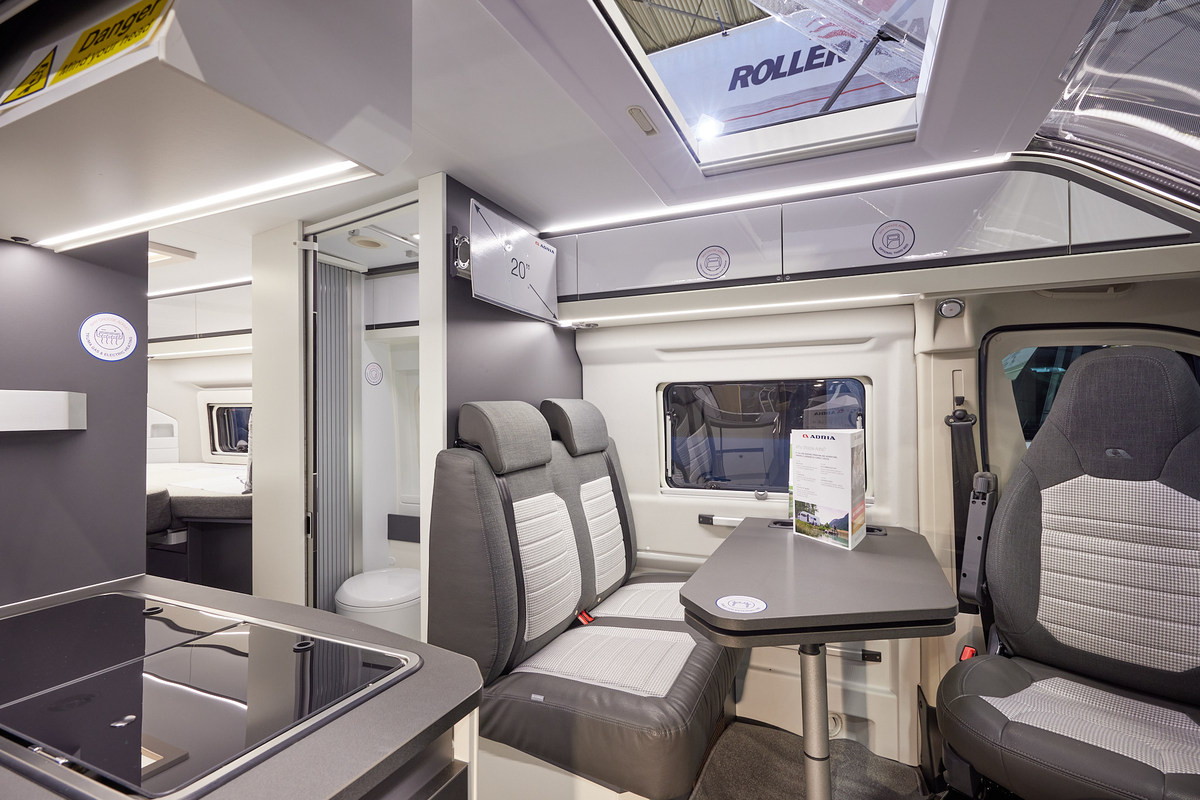 Adria Twin Supreme 640 SLB interior, rear passenger seats in brown and cream, fixed table, swivelled driver’s seat which can double as a dining chair.  Also shown is a hob, an open roof light and the combined washroom and toilet.  