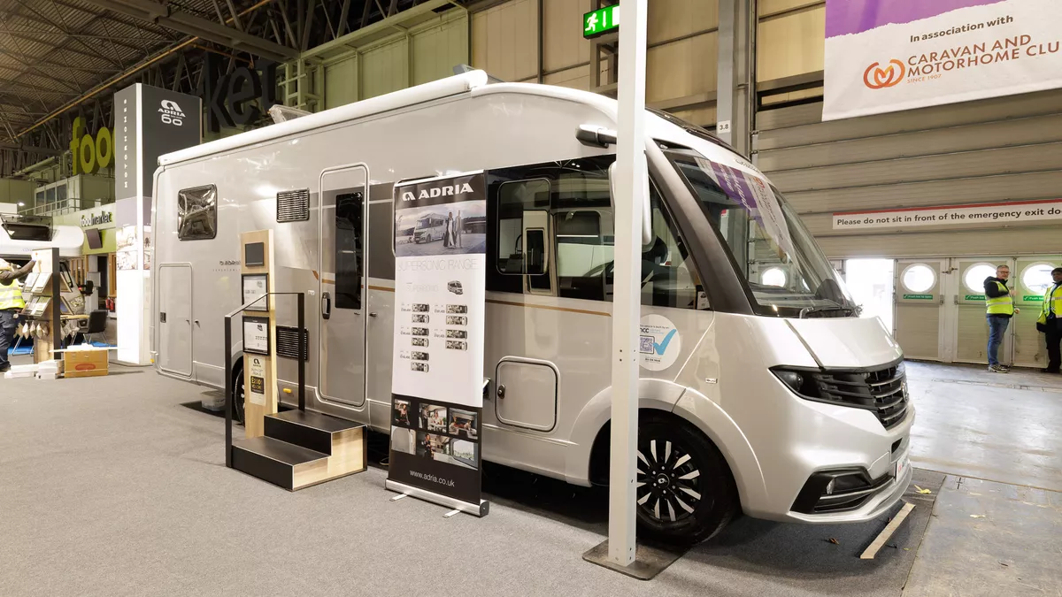 The Adria Supersonic 780 DL is silver.  There are two steps beside its habitation door.  There is a roller banner one side of the steps with an interaction stand on the other side.  Its skylights are open.
