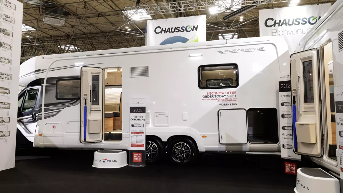 The Auto-Trail Frontier Comanche has a grey cab with matching decals.  Its habitation door is open and has a step to gain access.  There is a blue Auto-Trail umbrella in the door.  The garage door is open.