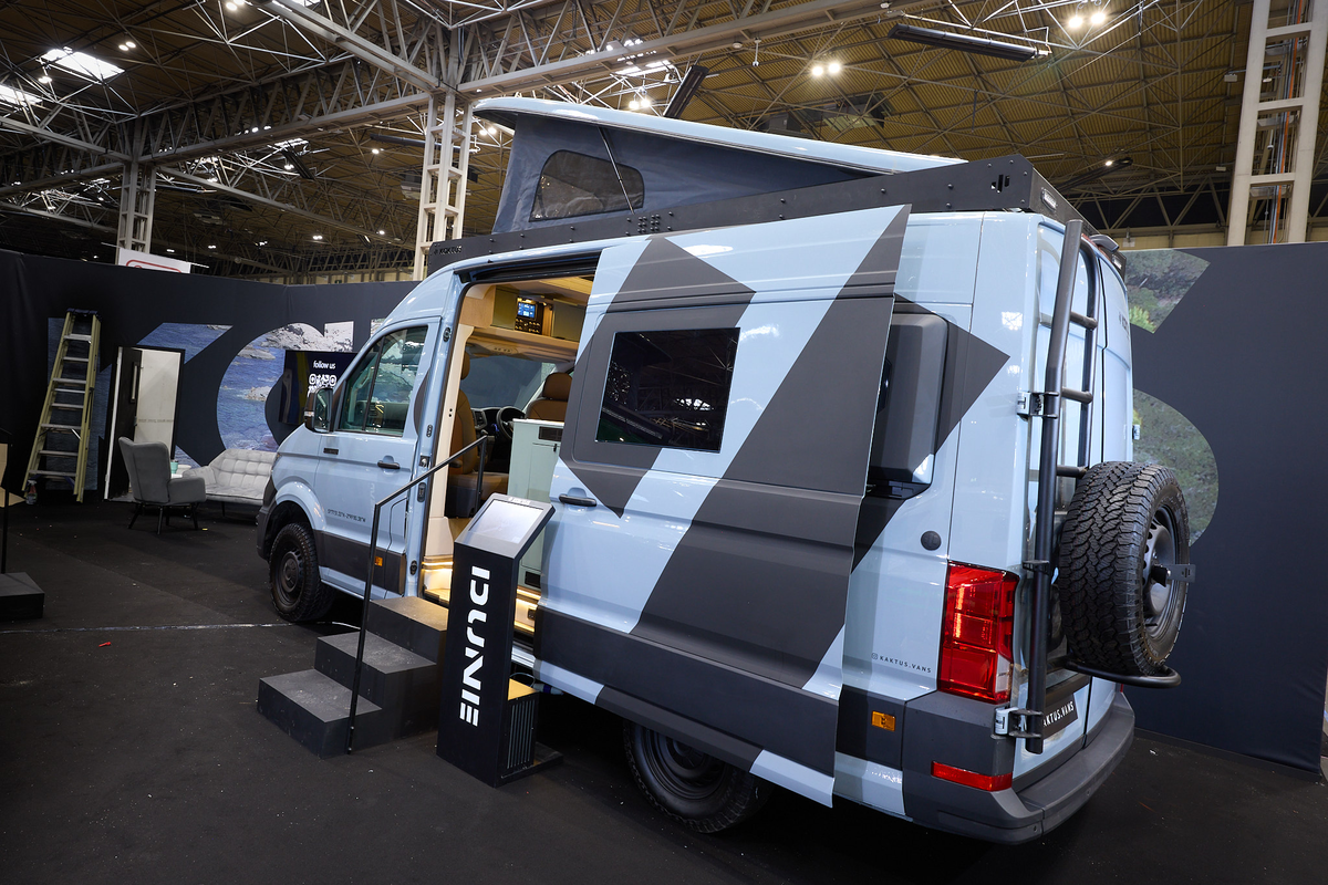 The Kaktus Adventure Vans Dune x4 has a pale blue and dark grey exterior with prominent decals.  There are two steps beside the open sliding door.  The pop top roof is fully extended.