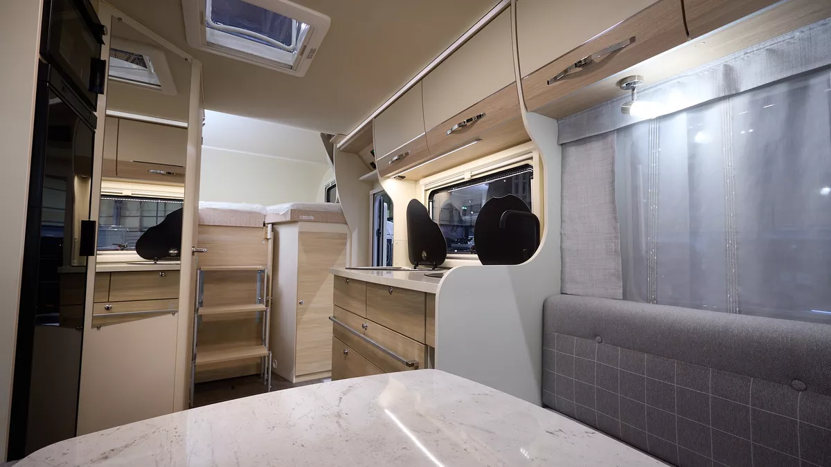 Eura Mobil Activa One 650 HS has a large white table and its upholstery is grey with white chequer pattern.  The cabinets are cream and pale wood.  There are three steps to the fixed rear bed.
