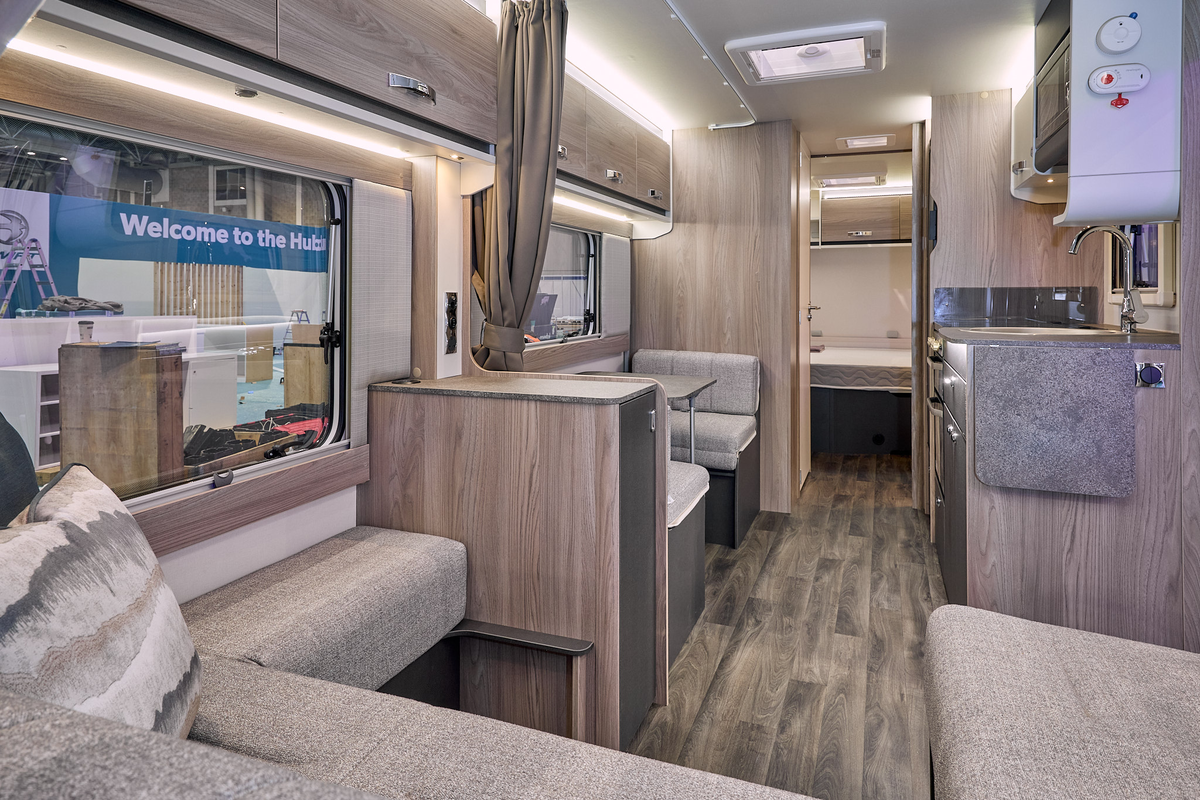 Swift Sprite Exclusive Quattro MB’s interior has a beige U shaped sofa with wooden furniture, there is a dinette opposite the kitchen unit.  The rear door is open revealing the fixed island bed.  