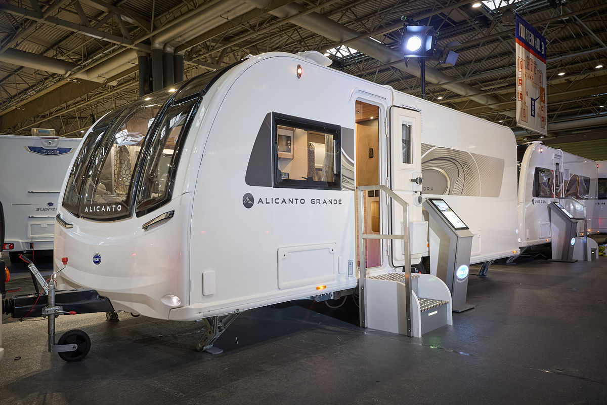 Bailey Alicanto Evora’s exterior, white with a large front window.  The entrance door is open and there are fixed metal steps into its interior.  There is an interactive stand next to the door.