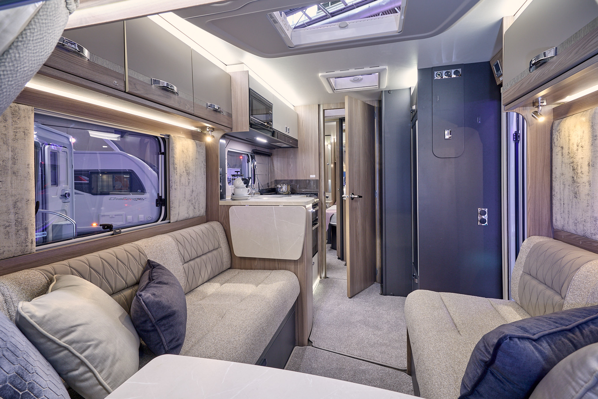 Swift Challenger Exclusive 560’s interior has wooden furniture with dark locker doors and beige upholstery which has embossed patterns.  There are three cushions on each sofa.  The rear door is open revealing the bedroom