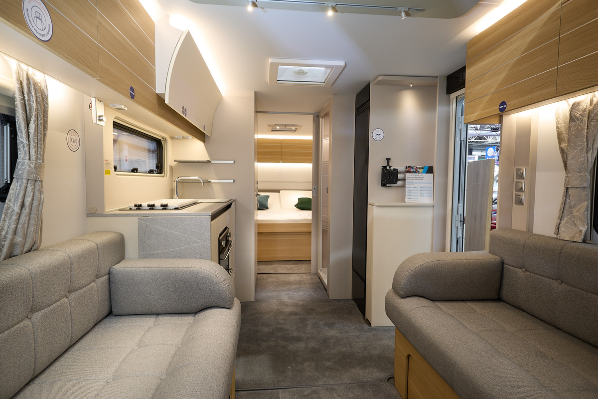 Adria Adora Tiber’s interior, it has wooden cabinets with grey upholstery in the lounge.  The walls and ceiling are white.  The kitchen has a domestic style oven and hob. There is a large fixed bed at the rear.
