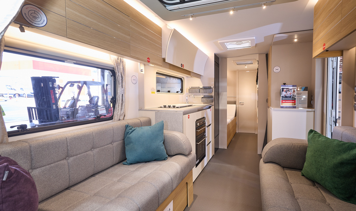 Adria Adora Seine’s interior, it has wooden cabinets with grey upholstery in the lounge.  There are three cushions, blue, green and purple.  The kitchen has a domestic style oven and hob.  The view leads to the bedroom with 3 skylights. 