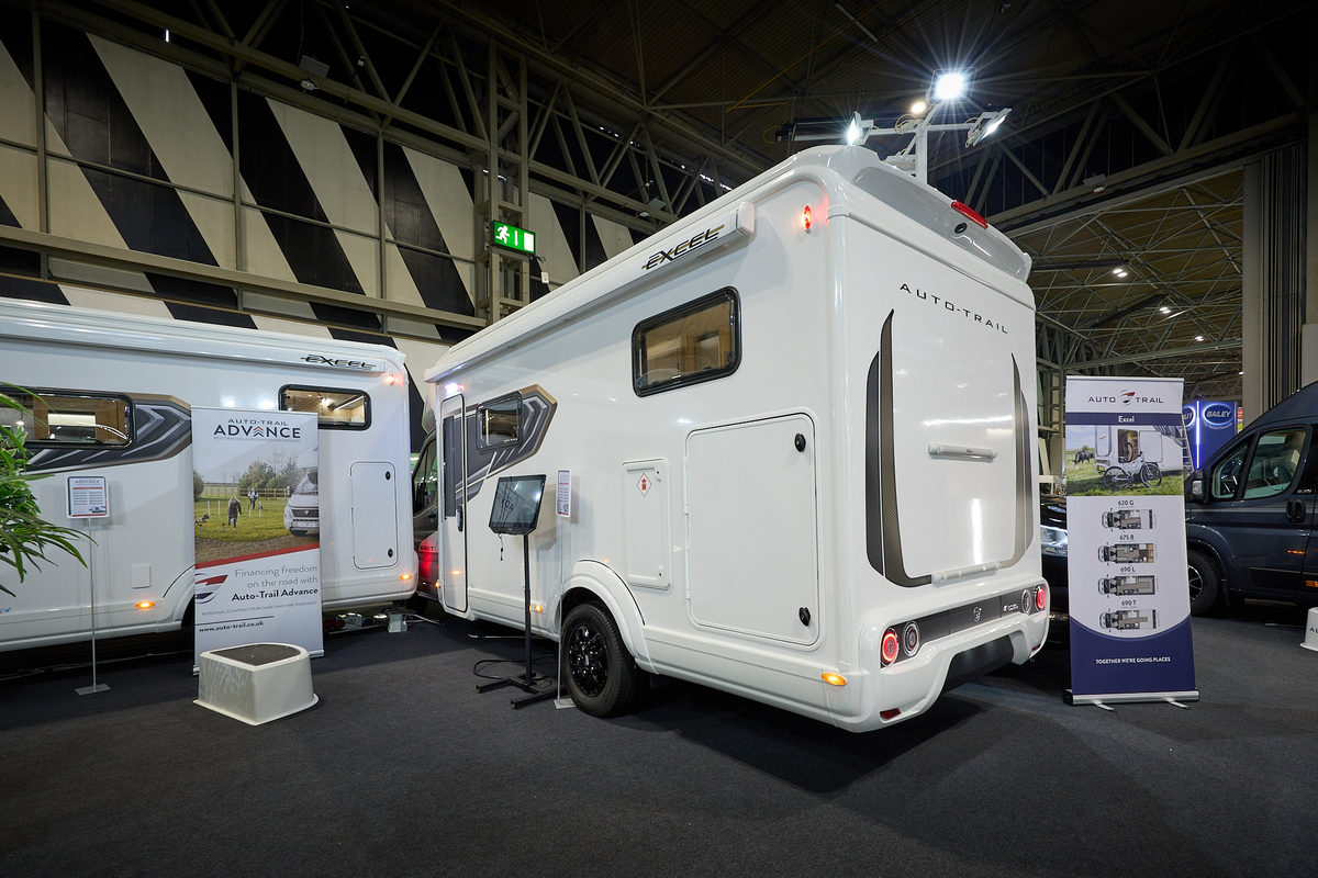 Auto-Trail Excel 690T’s exterior is white with dark grey decals.  It has a large door to the rear for its garage.  There is an interactive stand next to the rear wheel.  There is a roller banner at the end of the vehicle.