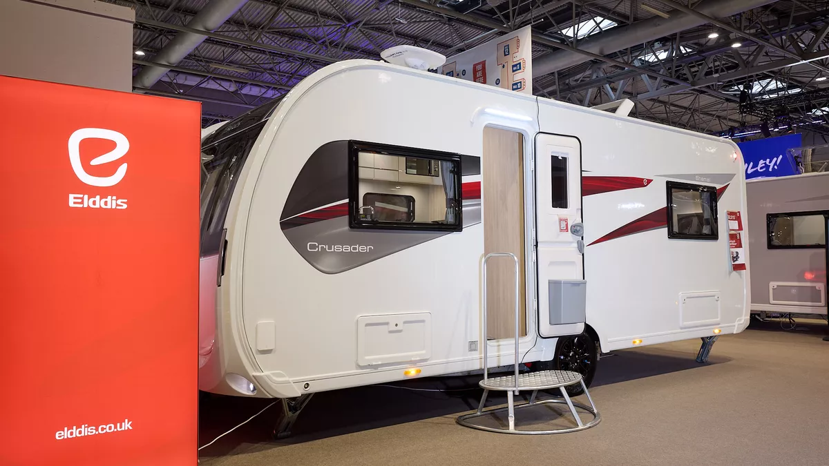 The Elddis Crusader Shamal has a white exterior with grey, red and black decals.  The entrance door is open and there is a metal step to gain easy access.  There is a large Elddis red banner with their website address next to it.