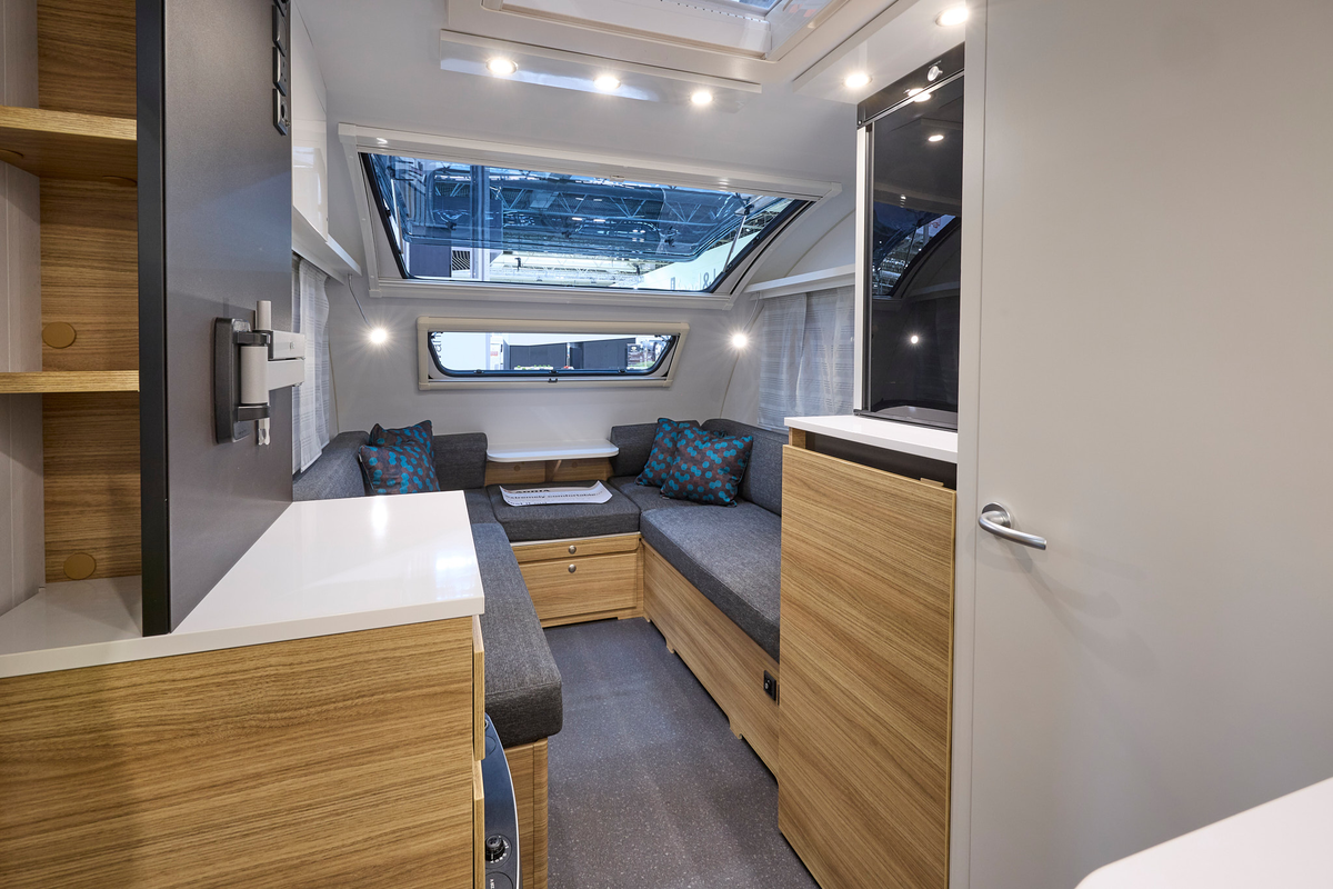 Adria Action’s interior, it has a white door, walls and ceiling, with wooden furniture and white surfaces. The sofas have dark grey upholstery with blue/grey patterned cushions and the floor is dark grey. It has a large panoramic window.