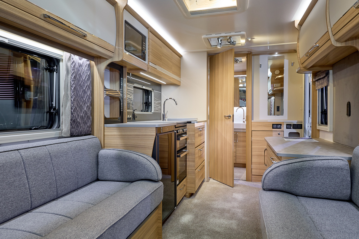 Bailey Unicorn Seville interior, it is grey with light wood.  The kitchen has a full oven and an under the counter fridge.  There is a rear washroom beyond the kitchen and its door is open.  
