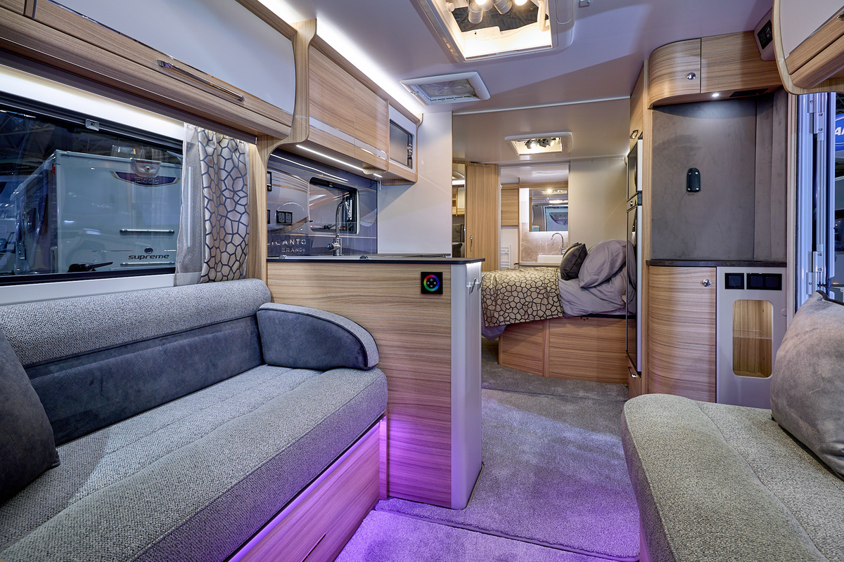 Bailey Alicanto Evora’s interior, it is grey with light wood.  The upholstery is grey and there is purple accent lighting.  The fixed island bed is beyond the kitchen and the washroom door at the rear is open.
