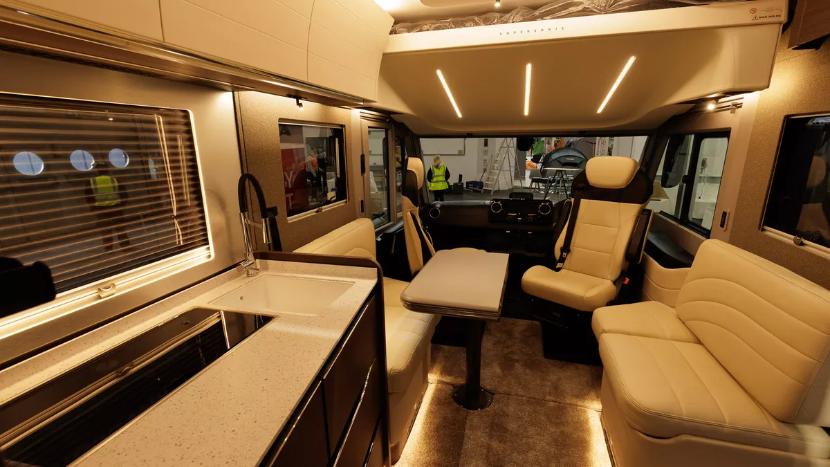 The Adria Supersonic 780 DL has cream upholstery and the front seats are swivelled.  There is a table which is folded but can be extended.  There is a roof bed above the cab.  The kitchen is to the left.