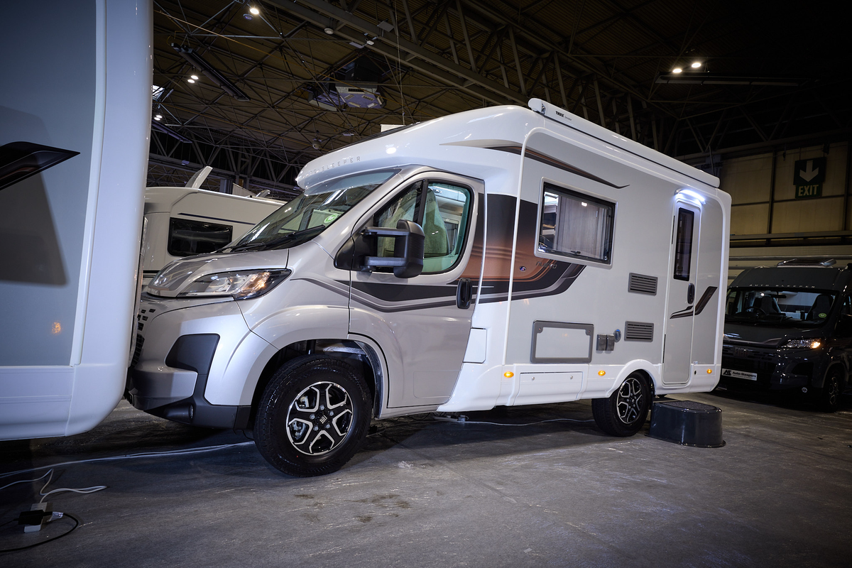 The Auto-Sleeper Nuevo has a gold cab with a white exterior with a grey inset. It has black/grey decals.  There is a black step by the habitation door.