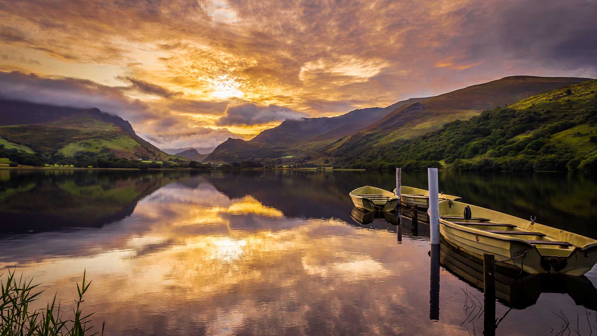 Looking out onto the waters of Llyn Nantlle Uchaf, three wooden boats are on the right leading to the mountains fading in the distance under a beautiful cloudy sunset sky all reflected in the calm waters