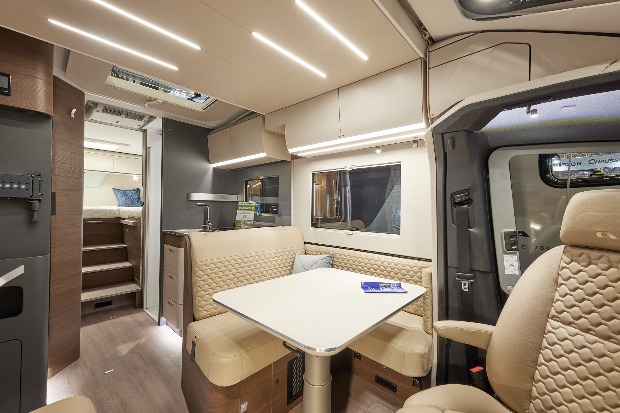 Adria Matrix Supreme MB 670 SL has cream embossed upholstery, with cream overhead cabinets and wooden curved panelling, it has mid-brown wooden flooring, there are three steps leading to the fixed rear bed with a blue cushion with lots of lighting.