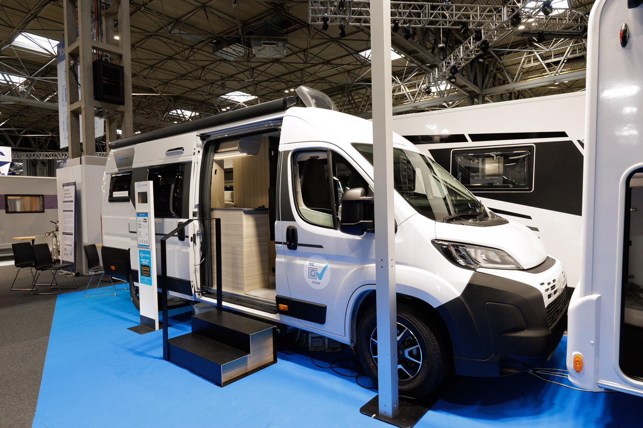 The Sun Living V Series 65 GX has a white exterior with black trim.  Its exterior door is open and there are two black steps to gain easy access.  There is a display stand next to the steps