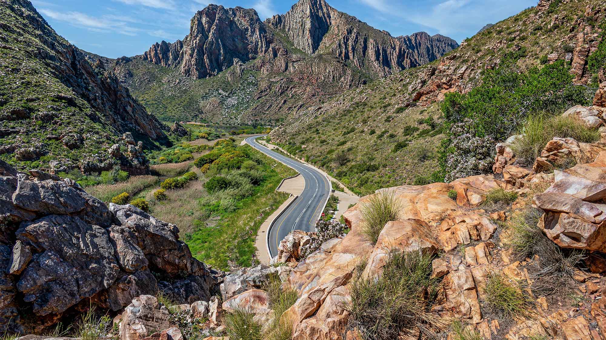 View from behine rocks of a road going through a hilly valley with a rocky mountain in the distance