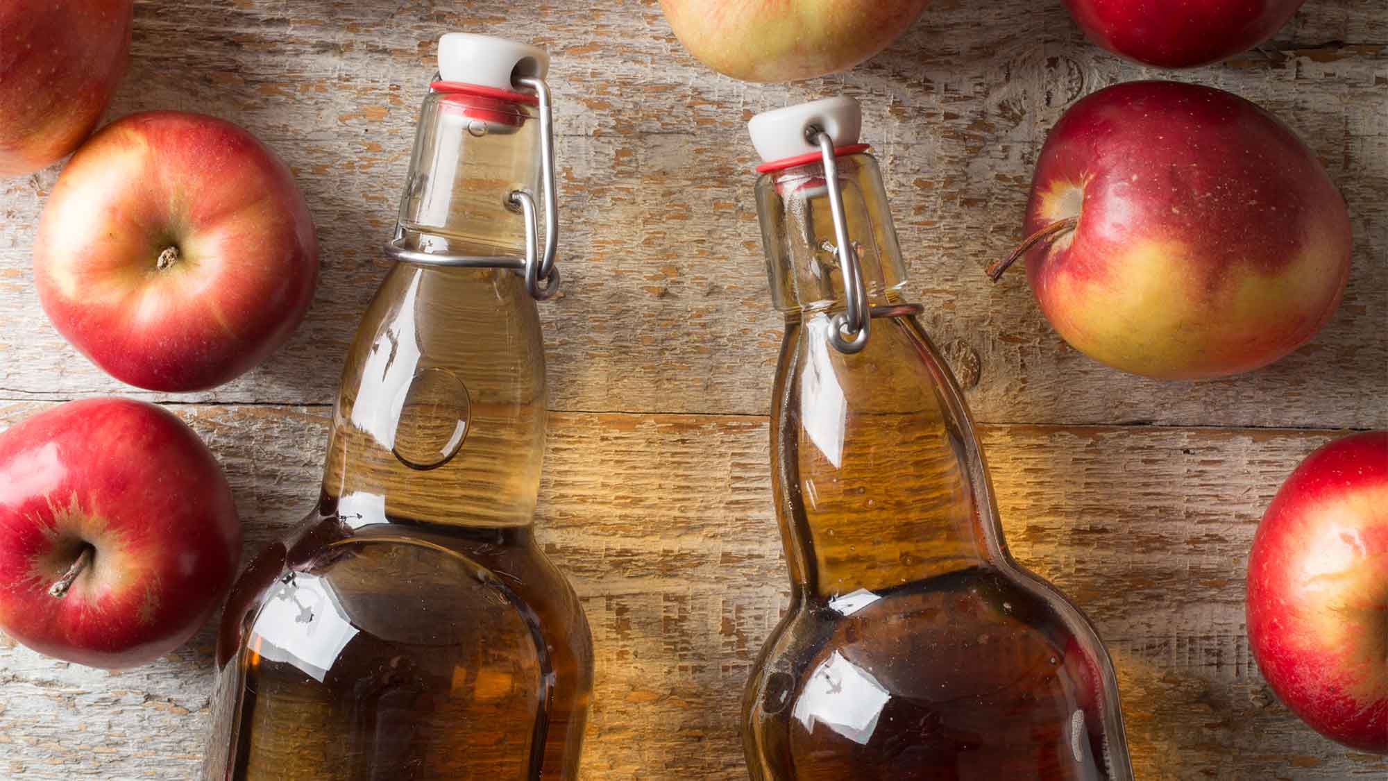 Two see through glass bottles filled with cider, laying on the table with two red apples laying by the outside of each bottle