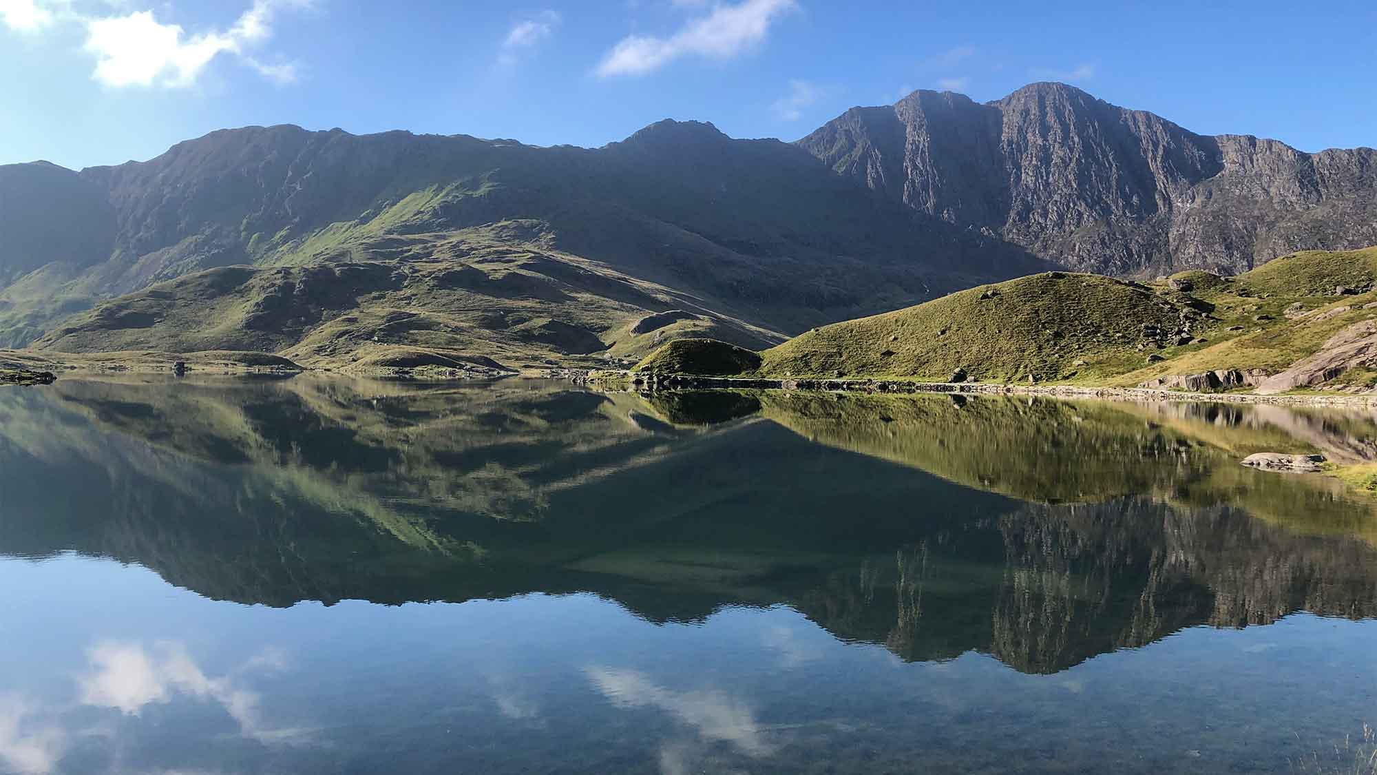 The mountain peaks rising high and wide with their reflections on the calm still waters of the lake on the Miners Track on Yr Wyddfa, Snowdon, under a bright blue sky.
