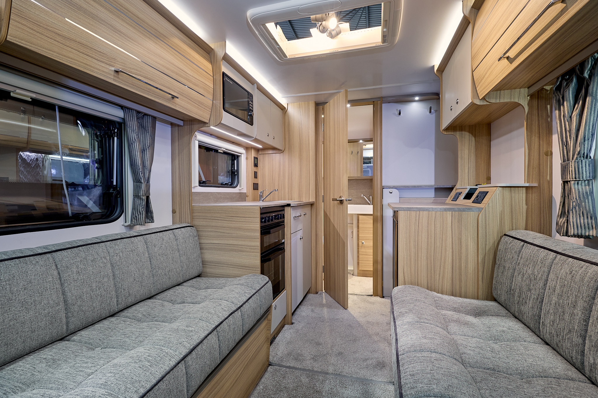 Bailey Phoenix GT75 420 has two grey sofas with a grey carpet.  The furniture is wooden and the rear interior door is open revealing the washroom