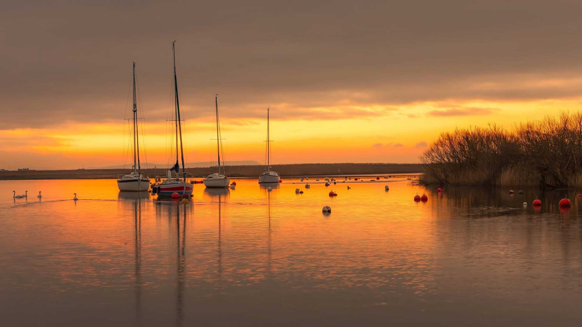 Four small boats on calm water with the orange sunset glowing in the sky and reflecting on the water