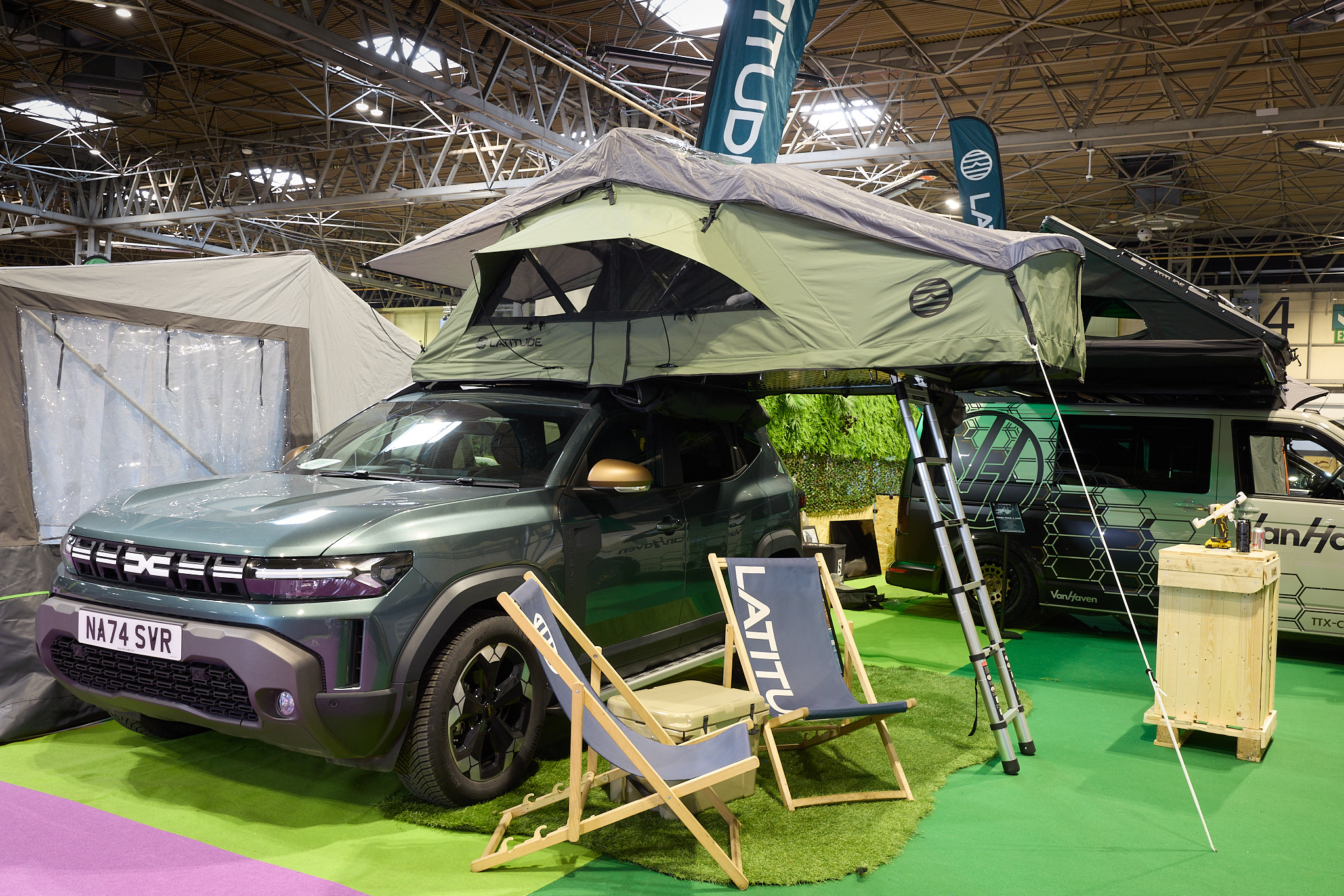 The Latitude Tents Pioneer is displayed on a green Dacia vehicle.  It has green canvas walls and a pale grey roof.  It has an extended platform with silver ladder to gain easy access.  There are two grey deck chairs by the Dacia.  