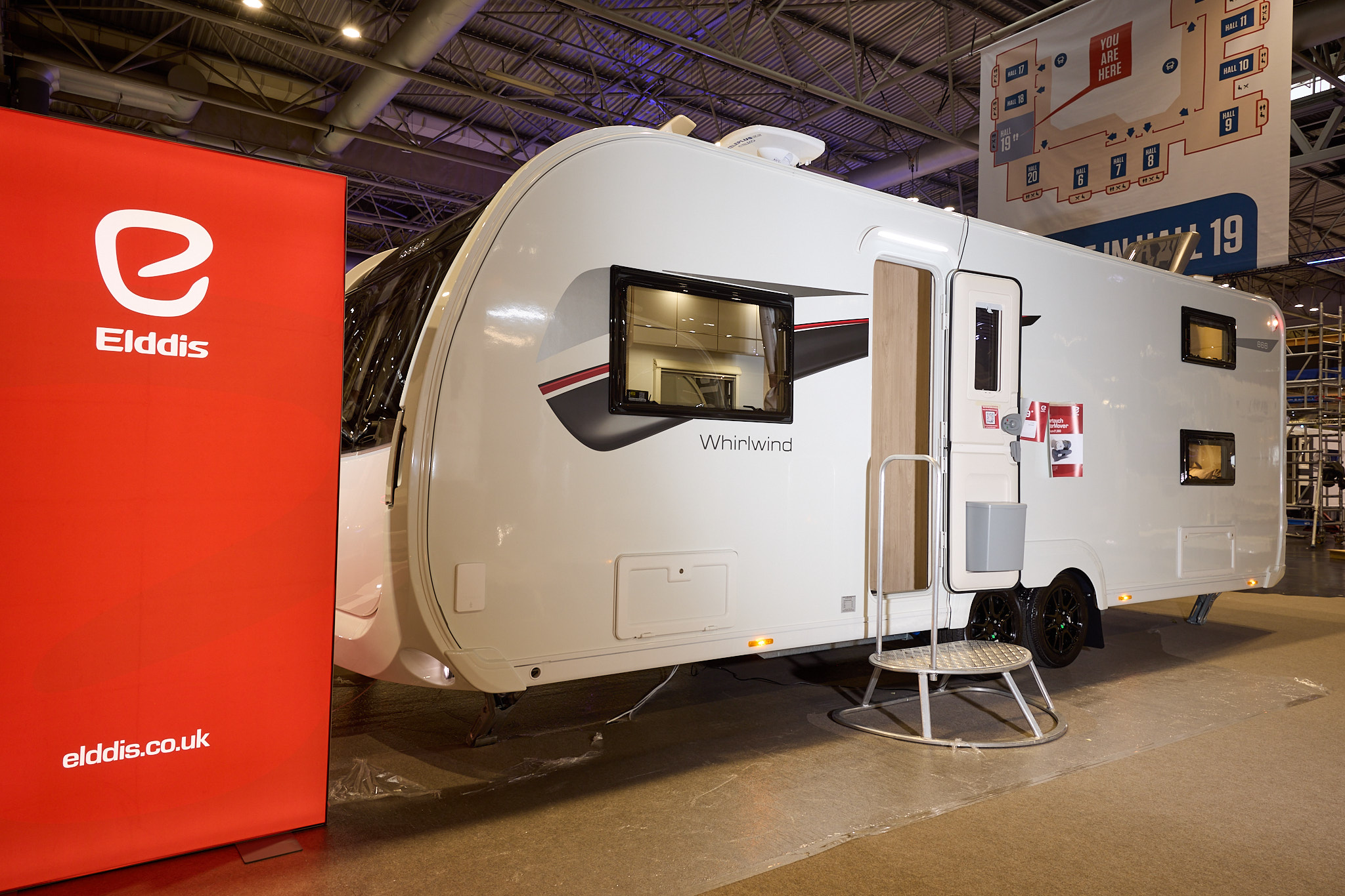 The Elddis Whirlwind Evolve 868 has a white exterior with a grey, red and black decal.  The entrance door is open and there is a metal step to gain easy access.  There is a large Elddis red banner with their website address next to it.