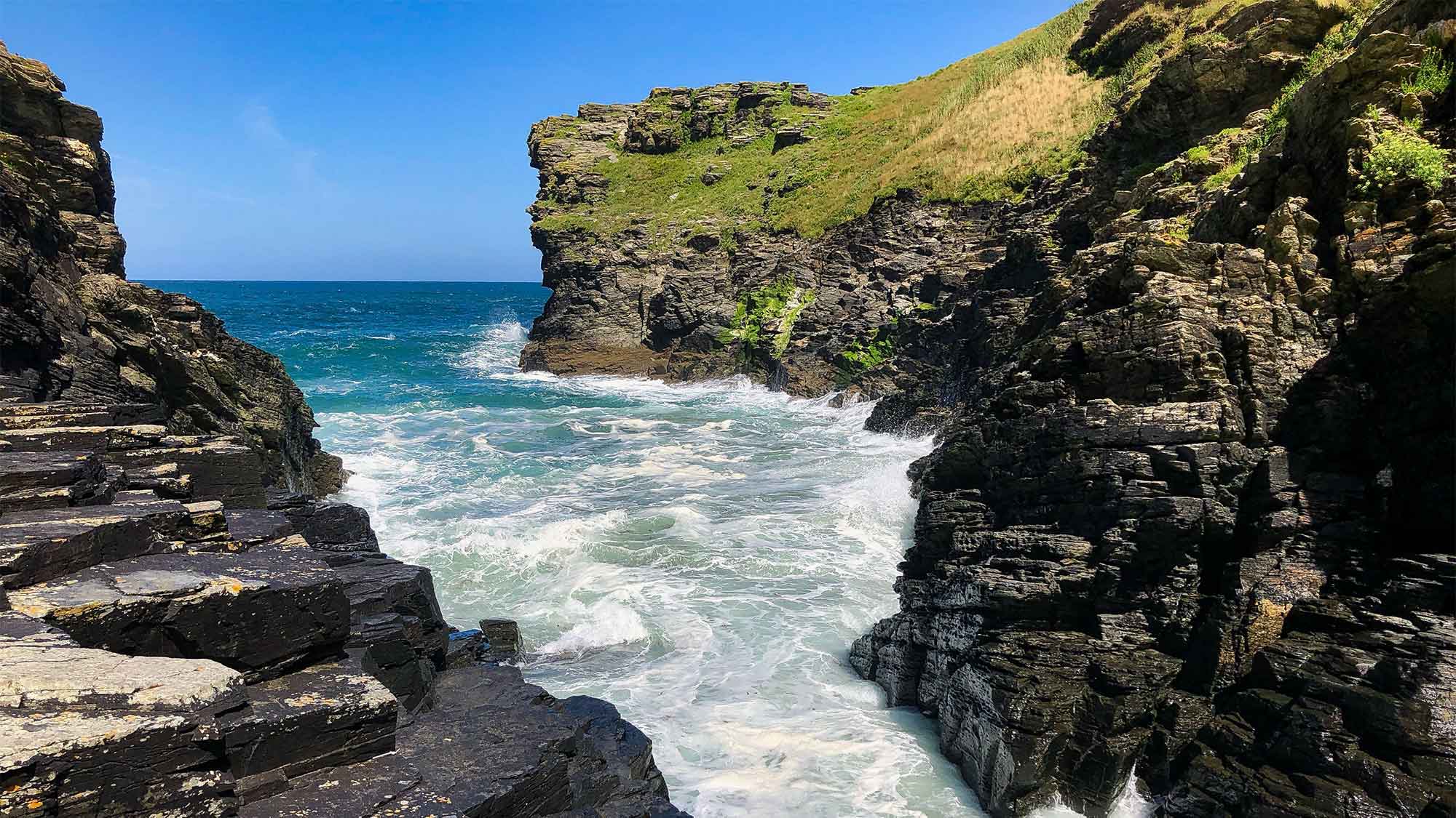 White water rushing in from the blue sea between rocky walls with a back drop of a clear blue sky meeting the sea. 