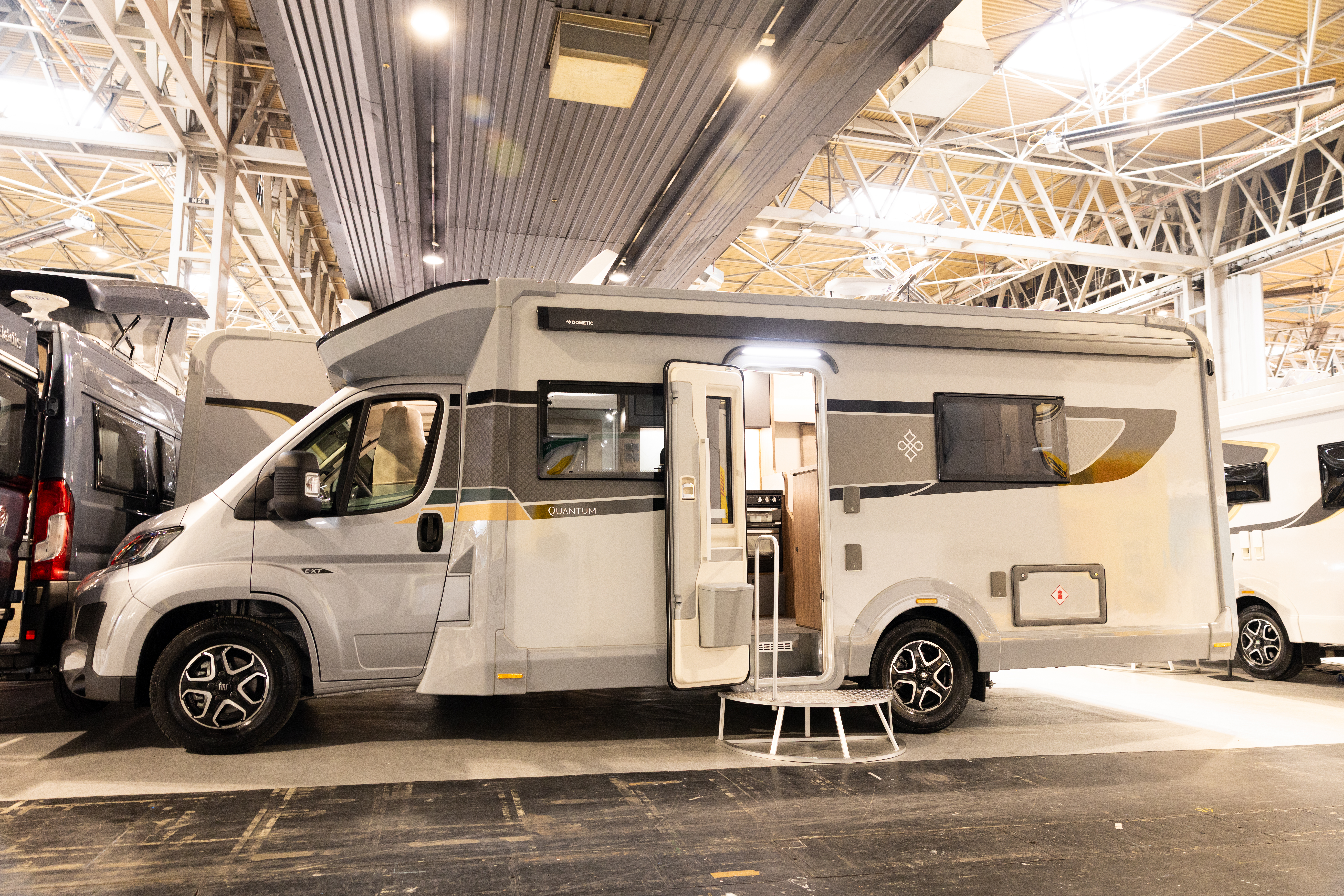 The Buccaneer Quantum F-4S has a silver cab with a white coachbuilt body.  It is decorated with decals of various colours.  Its habitation door is open and there is step to gain easy access.