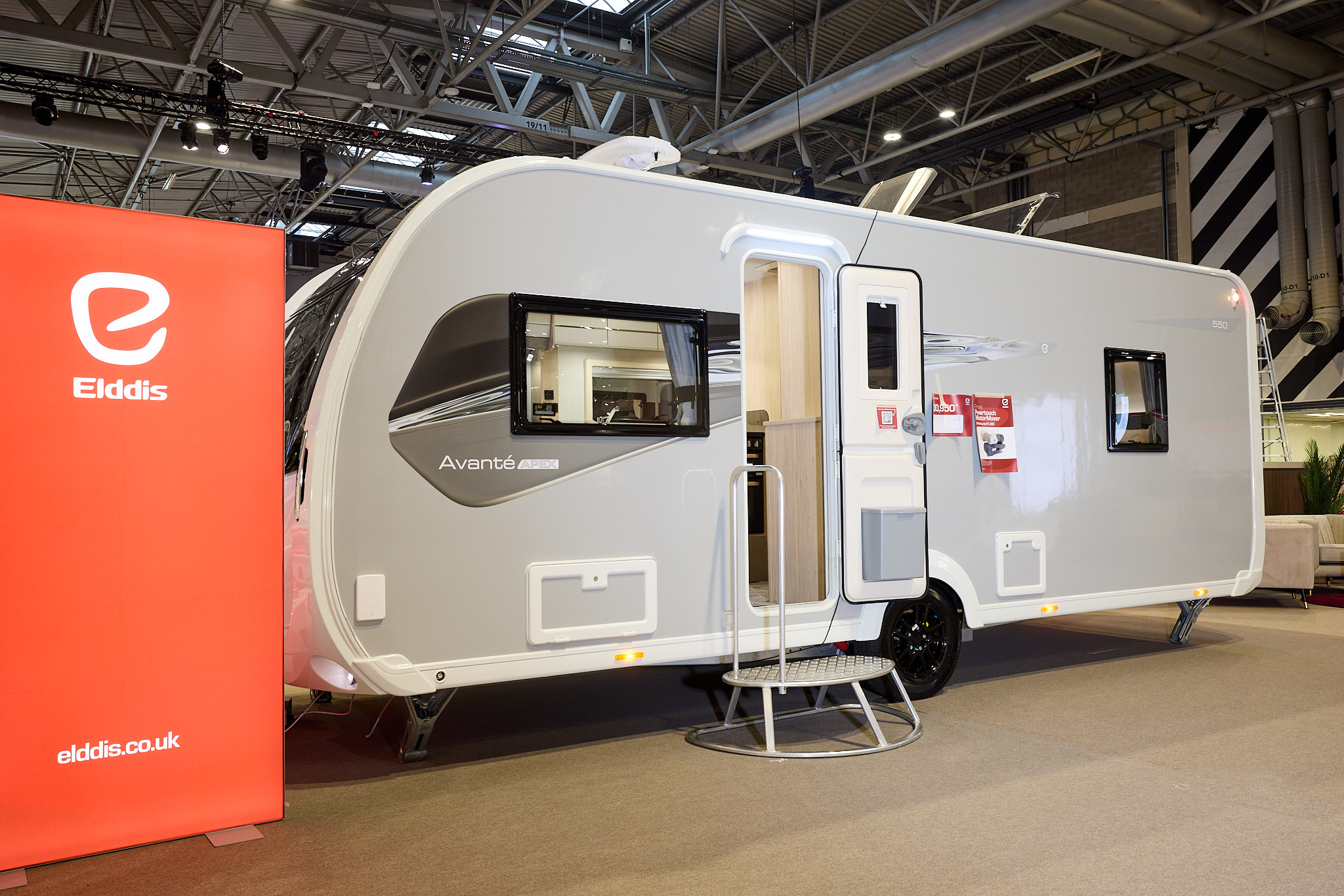 The Elddis Avanté APEX 550 has a grey exterior with a grey and black decal.  The entrance door is open and there is a metal step to gain easy access.  There is a large Elddis red banner with their website address next to it.