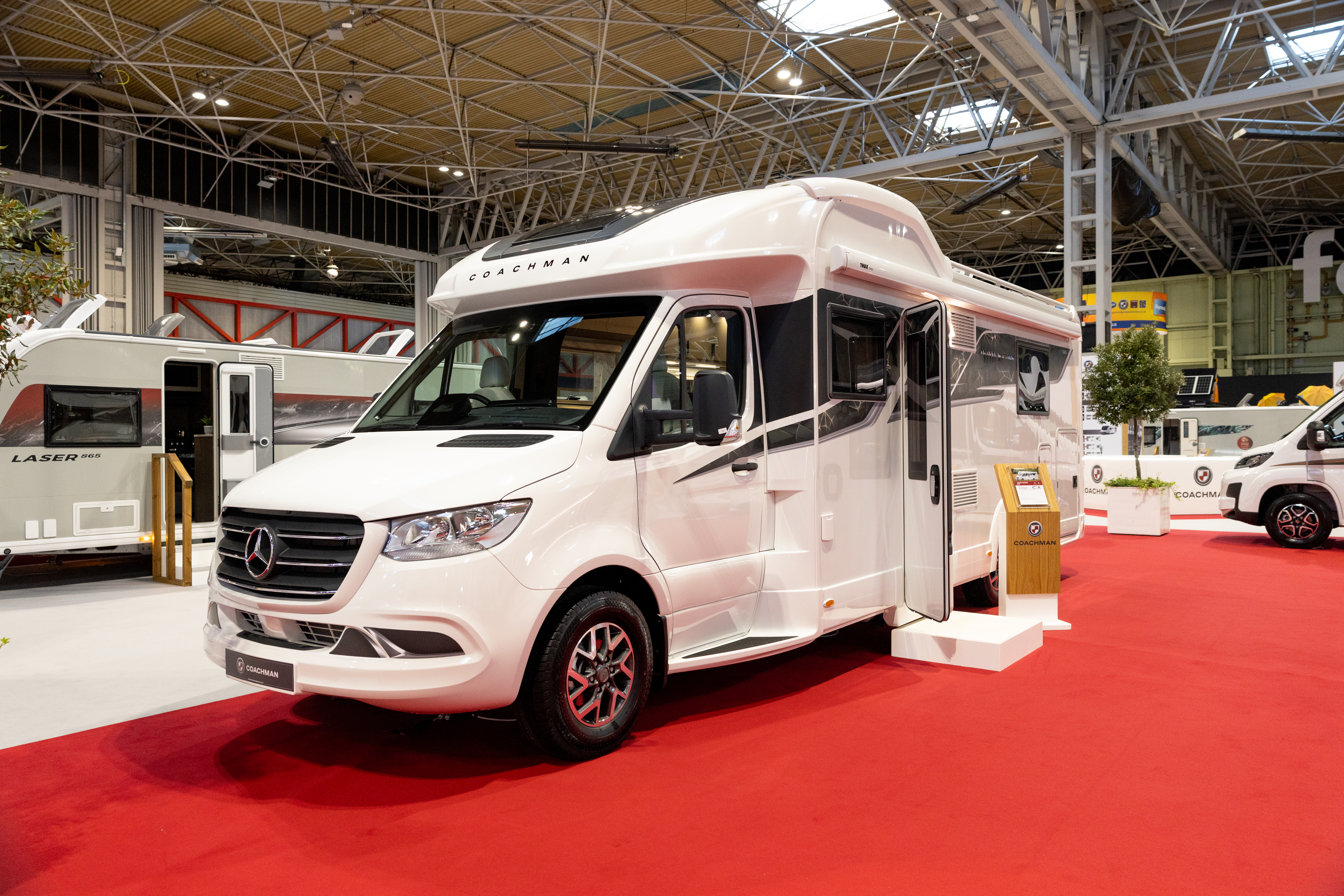 The Coachman Travel Master 545 is white, it is a Mercedes Benz.  Its habitation door is open and there is step to gain access.  Next to the step is a wooden stand