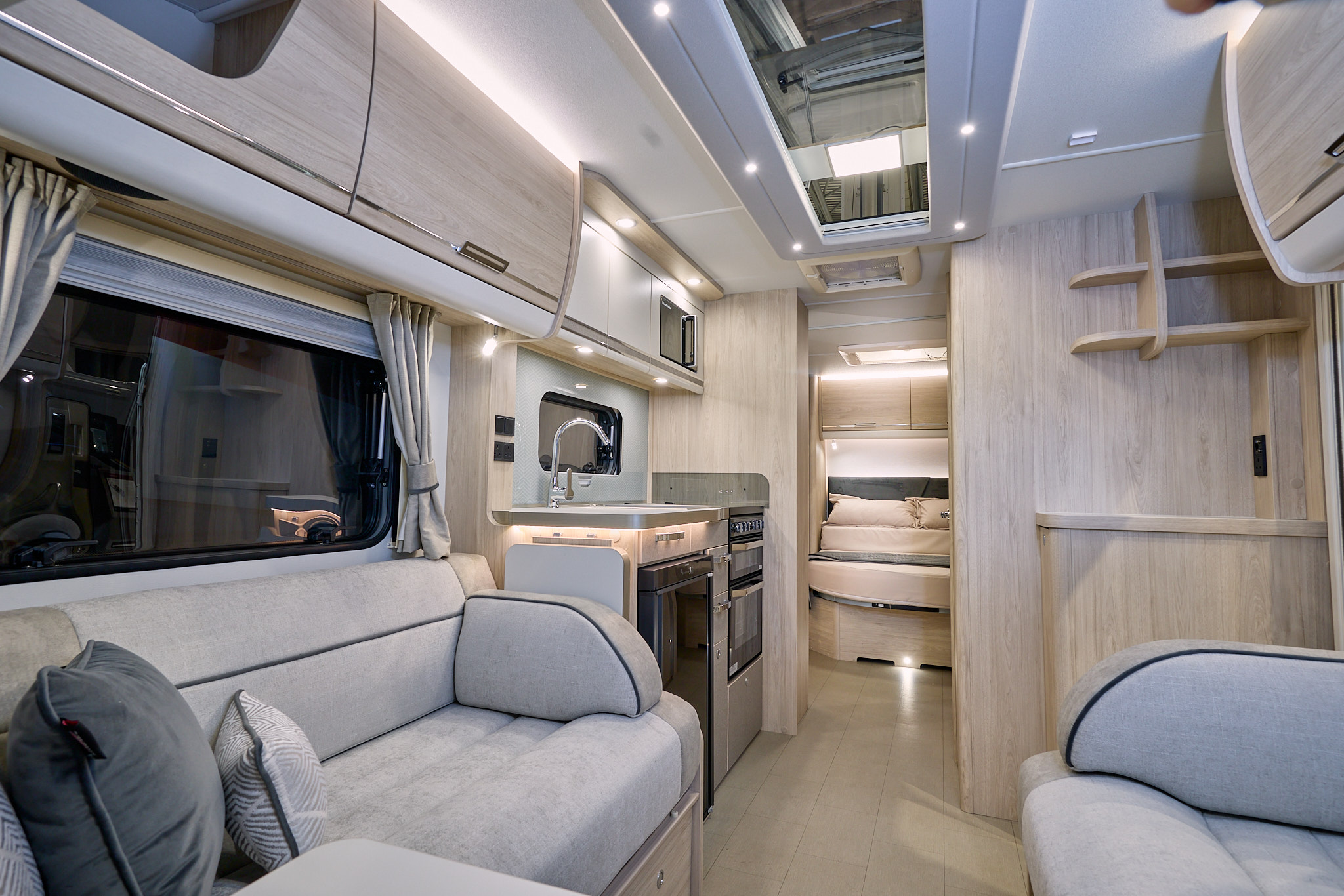 Elddis Crusader Aurora’s interior has grey upholstery with dark grey accents.  It has pale wooden furniture.  The kitchen is in the middle and the bedroom door is open revealing a fixed island bed.