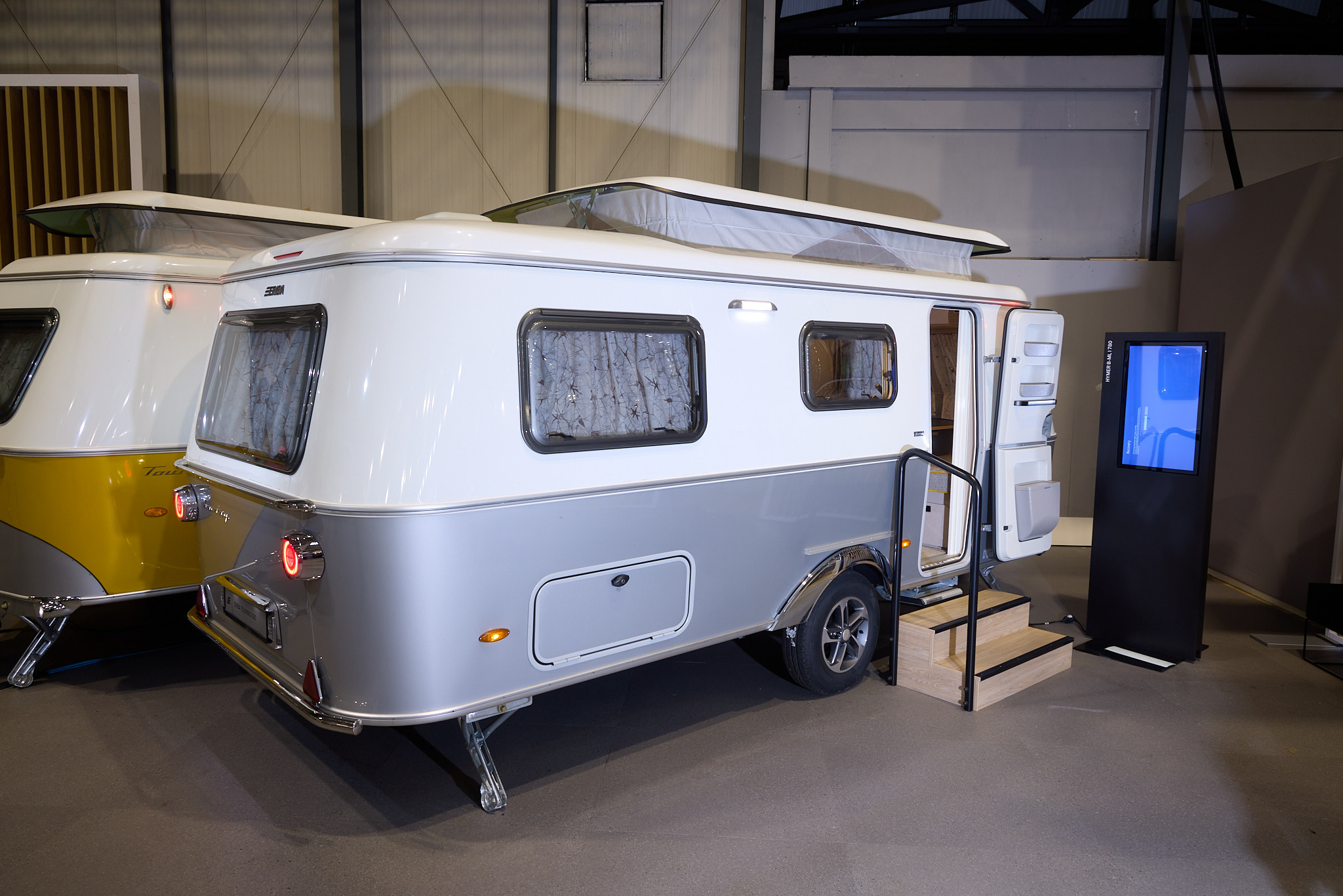 Eriba Touring 630 Urban’s exterior is silver and white.  It’s pop top roof is extended.  There are two wooden steps to gain easy access and there’s a large interactive screen to the right.