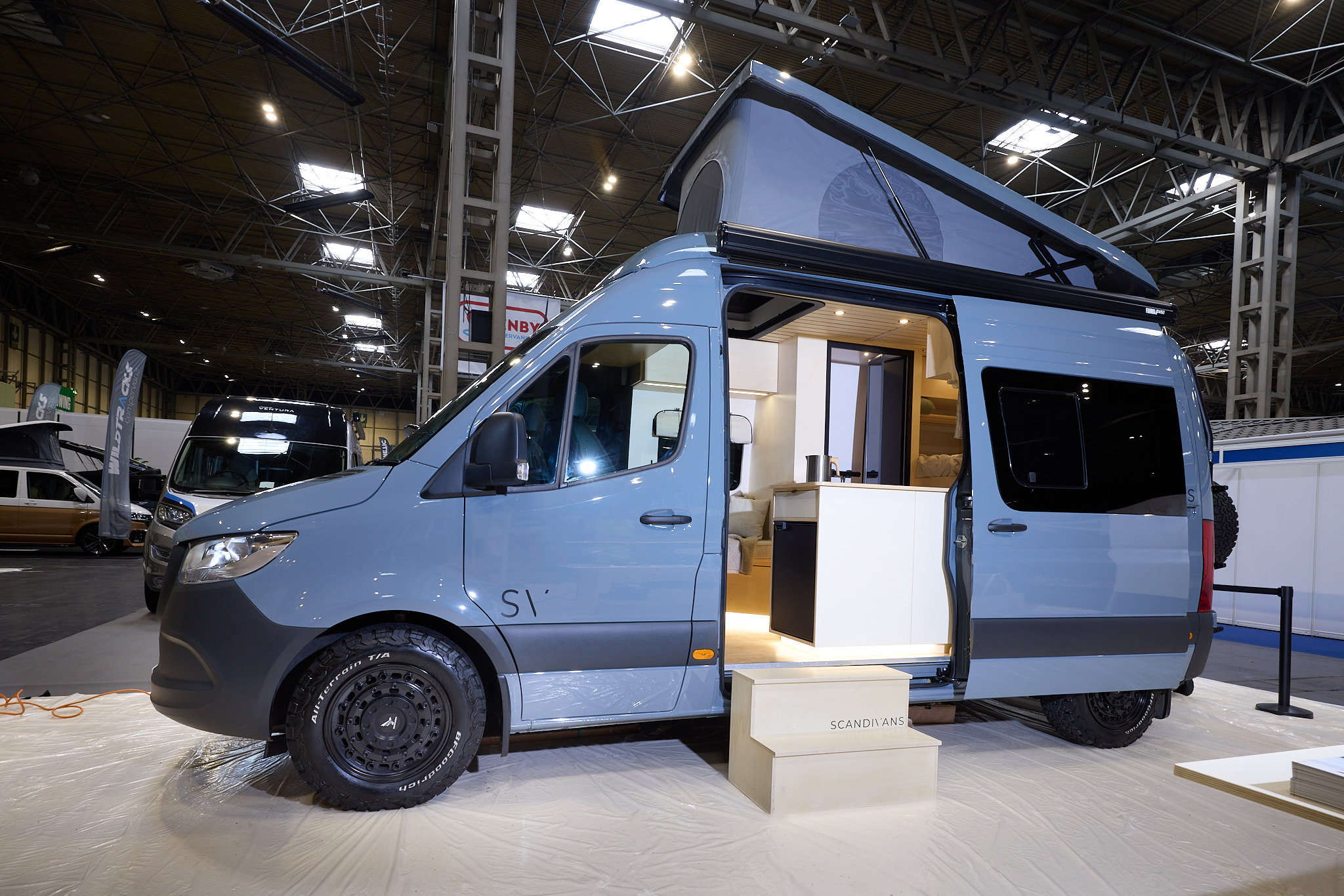 The Scandivans Sprinter 907 MWB L2H2 has a pale blue exteriors.  There are two steps leading to the open sliding door revealing the interior.  Its rising roof is fully extended.
