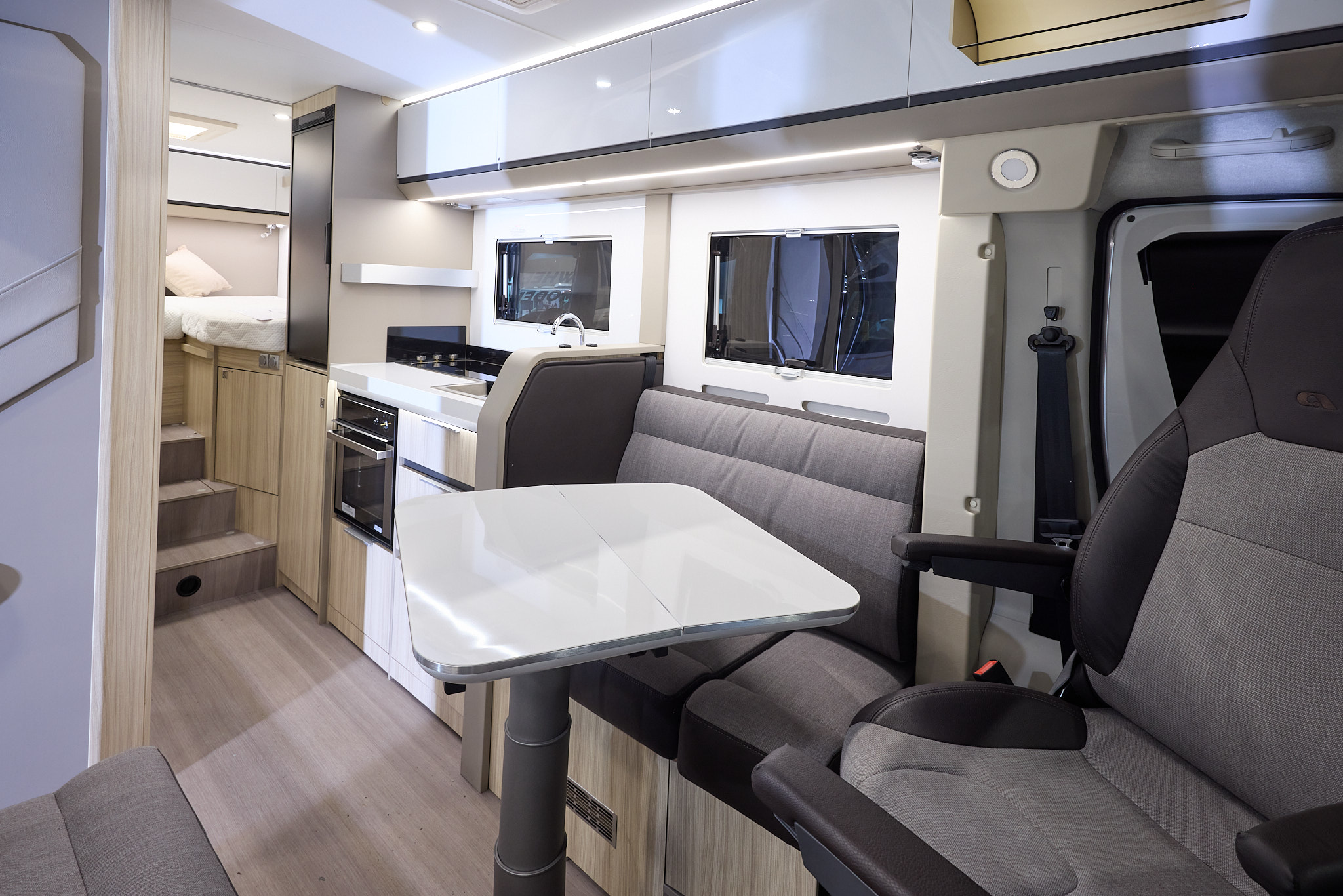 The Adria Compact Supreme has a wood style floor, with pale wood cabinets and white overhead lockers.  The upholstery is two tone with dark edging.  There is a white table which can be rotated.  There are three steps to the fixed raised rear bed.