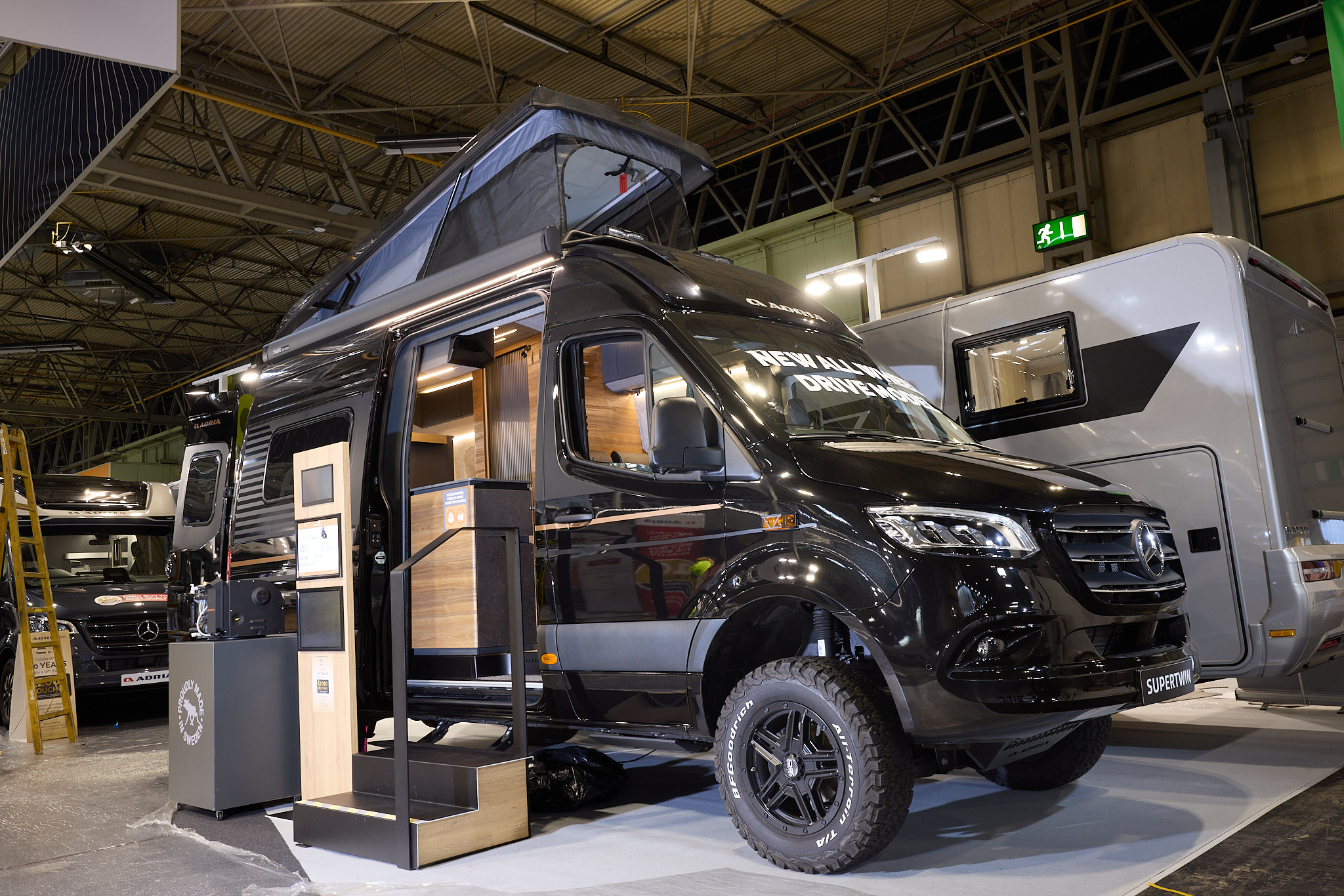 The Adria SuperTwin has a black exterior.  The habitation door is open and there are two steps to gain easy access. There is an information board to the left of the steps.  The rising roof is open and so are the rear doors.
