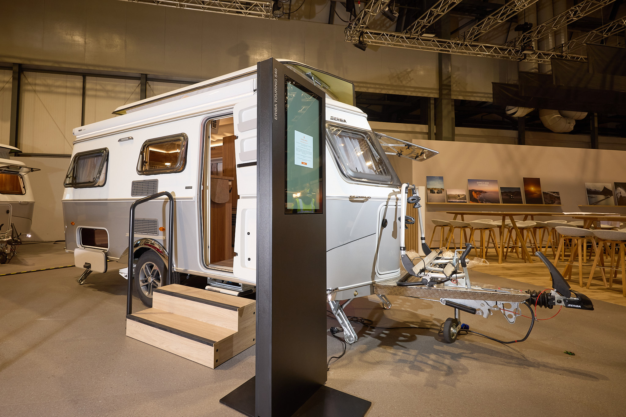 The classically styled Eriba Touring 540 Legend has a two tone layout in silver and white.  Its pop top roof is open as is its entrance door.  There are two wooden steps to gain easy access.