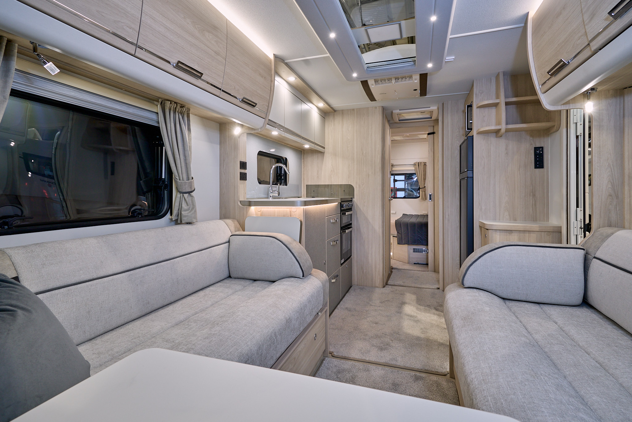 Elddis Crusader Zephyr’s interior has grey upholstery with dark grey accents.  It has pale wooden furniture.  The kitchen is in the middle and the bedroom door is open revealing a fixed island transverse bed.