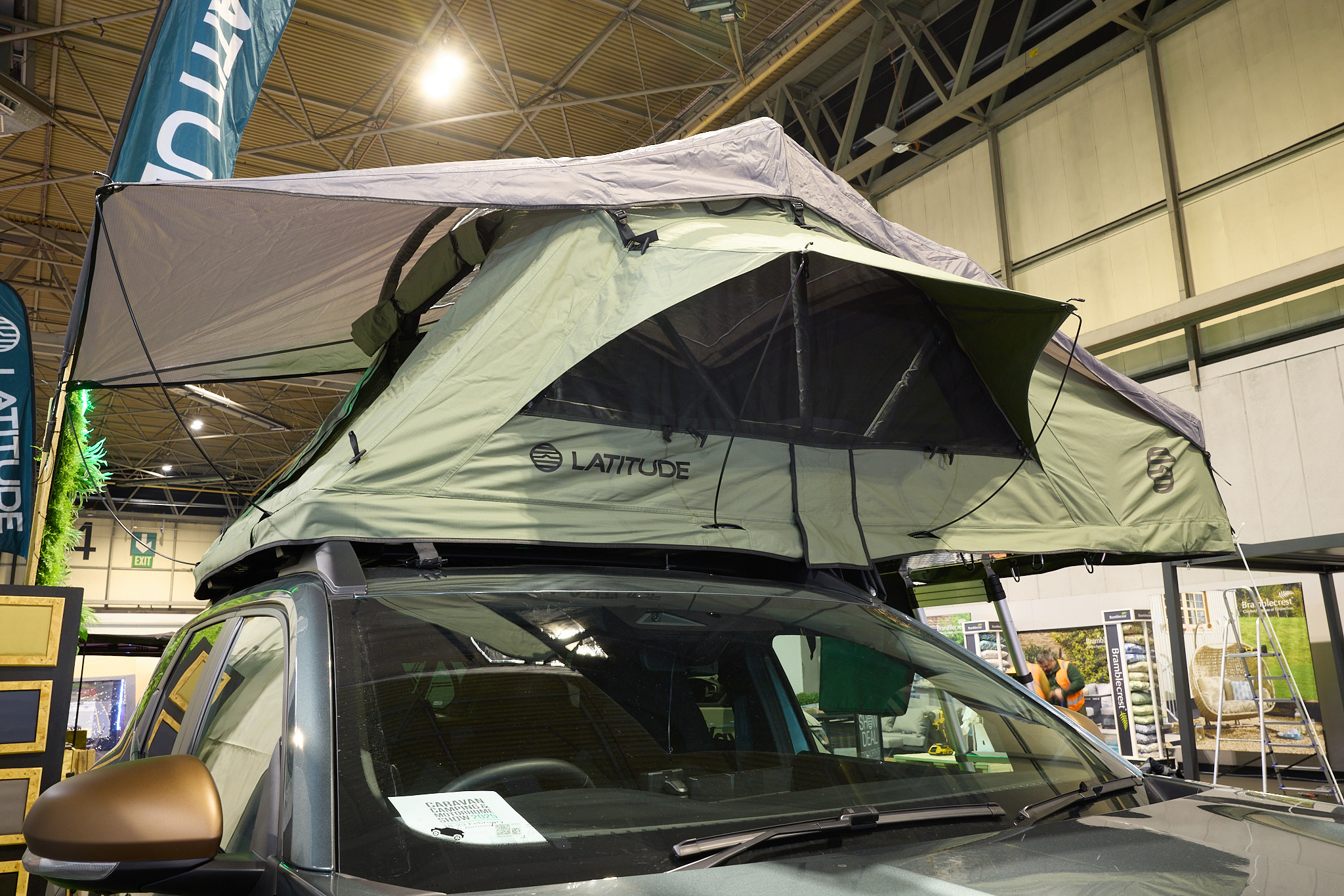 The Latitude Tents Pioneer has green canvas walls and a pale grey roof.  The window is open.  It has an extended platform with silver ladder to gain easy access