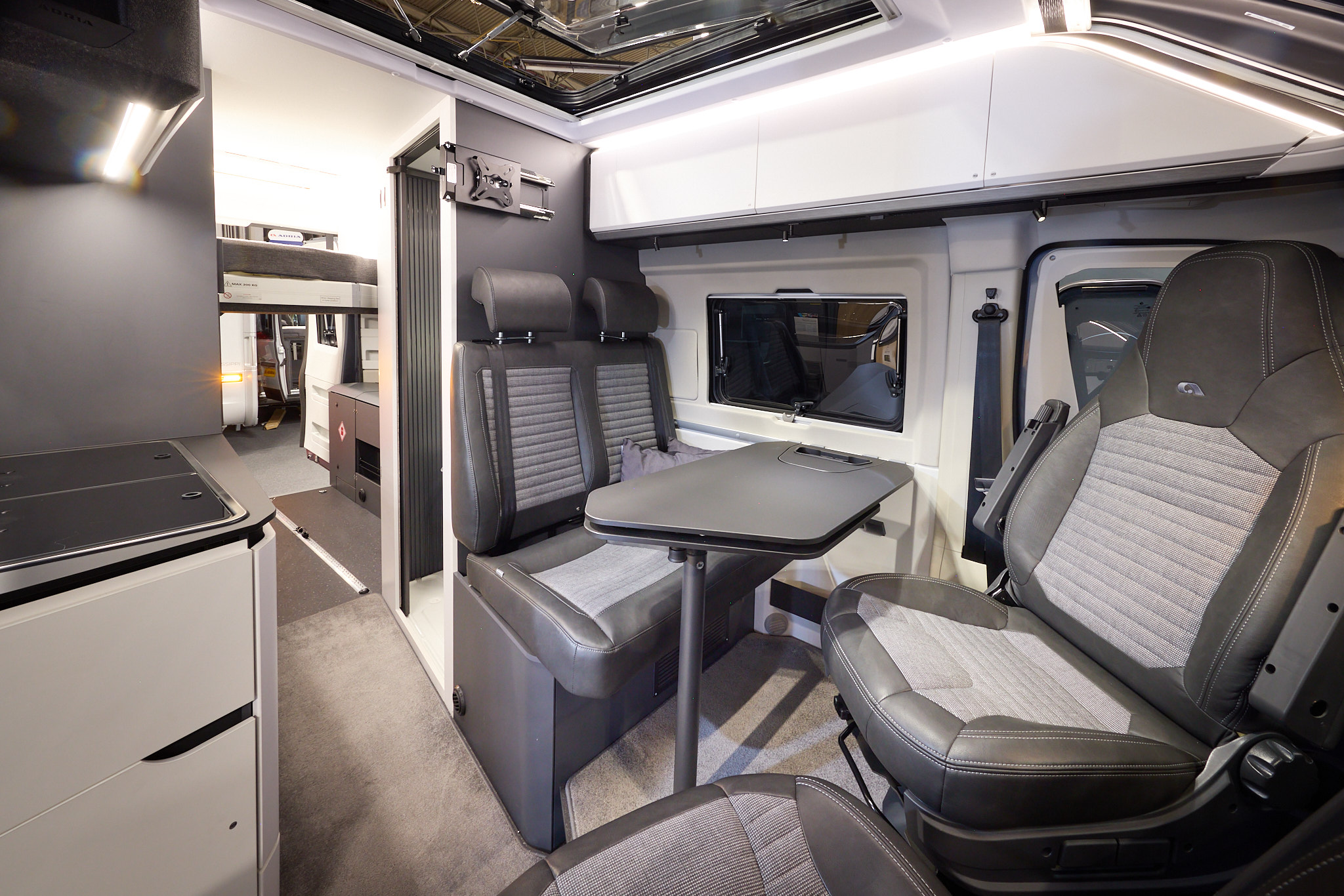 The Adria Twin Supreme’s floor has a pale brown carpet, with white cabinets and dark grey panels.  There is a large panoramic sunroof.  The upholstery is two tone with dark edging.  There is a grey table which can be rotated.  