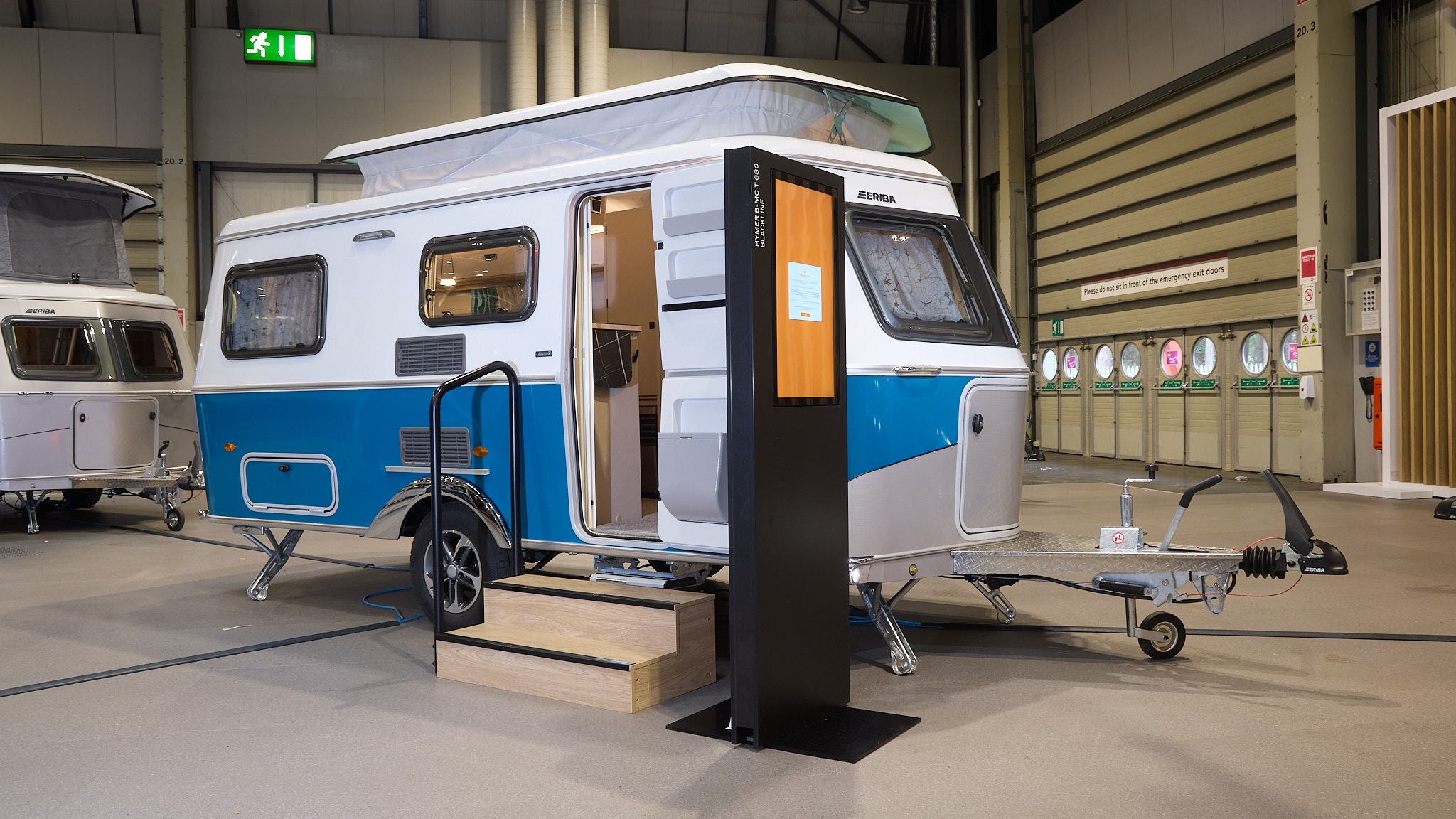 Eriba Touring 540 Harbour Blue Edition’s exterior is blue and white.  It’s pop top roof is extended.  There are two wooden steps to gain easy access and there’s a large interactive screen to the right.