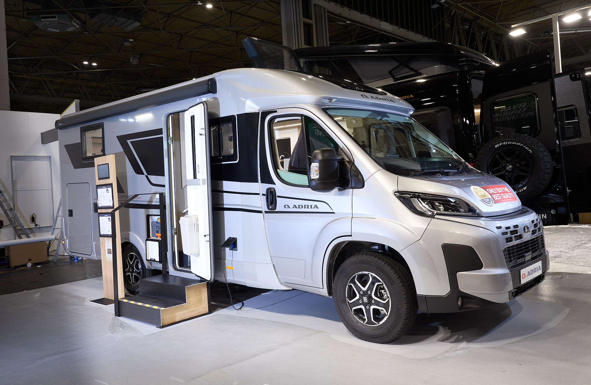 The Adria Compact Supreme has a silver exterior.  The habitation door is open and there are two steps to gain easy access. There is an information board to the left of the steps.  