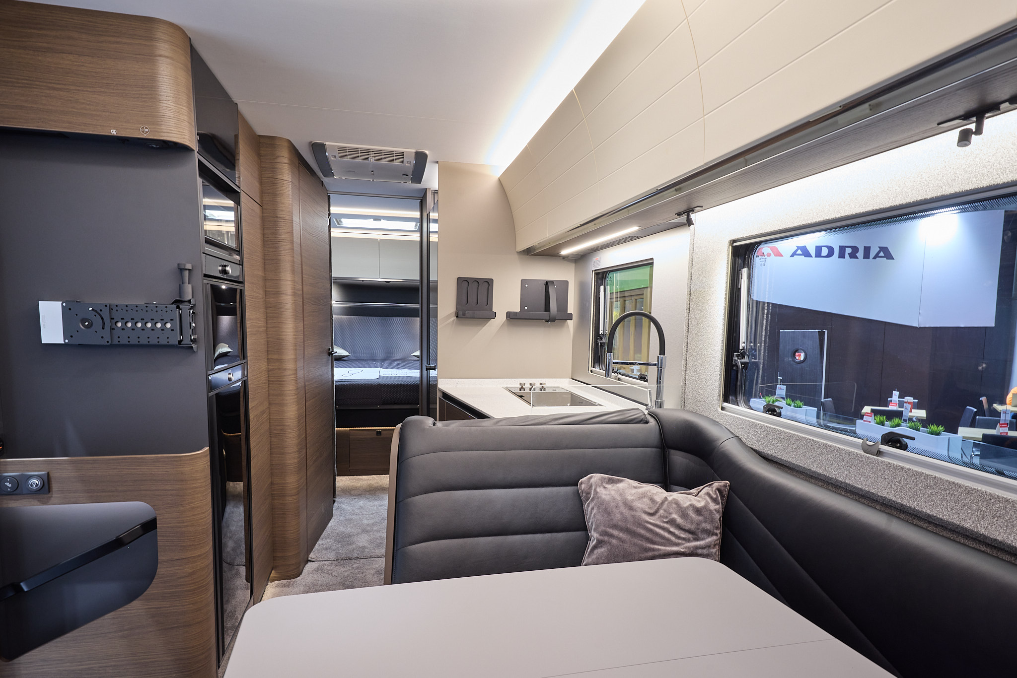 The Adria Supersonic’s floor has a grey carpet, with dark wood cabinets and cream curved overhead lockers.  The upholstery is dark leather.  There is a white table.  There is a fixed rear bed to the rear.