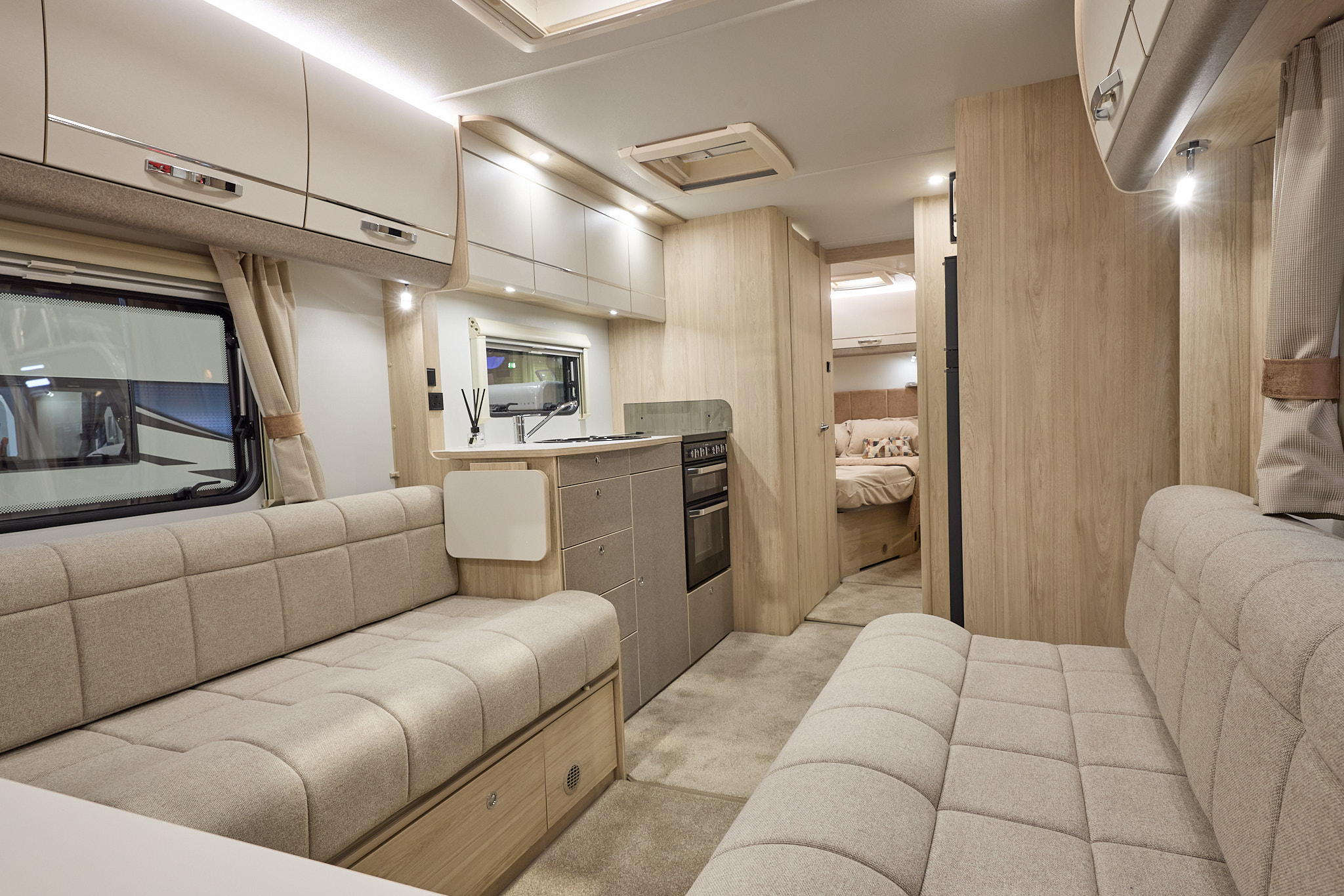 Interior of the Elddis Whirlwind Evolve 868 with cream sofas and wood accents and beige carpets.  The door to the rear bedroom is open.  Perfect for relaxation and designed for comfort.