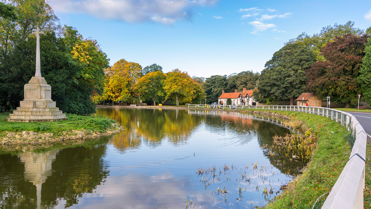 A river running alongside a road bending to the right with a Memorial Cross on a small rive island in Bishop Burton, East Riding